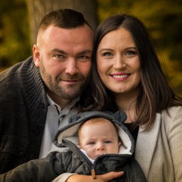 Oczami Zajawki Fotografia - Portret uśmiechniętej rodziny: ojciec z brodą, matka z długimi włosami i niemowlę w kurtce z kapturem na tle jesiennych drzew.