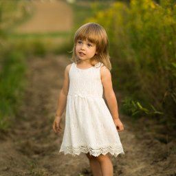 Oczami Zajawki Fotografia - Urocza, bosa dziewczynka w białej sukience stoi na polnej drodze, spoglądając w górę. W tle zieleń i rozmyty krajobraz. Delikatne, naturalne światło.