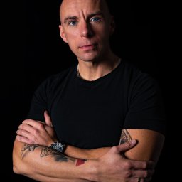 Oczami Zajawki Fotografia - Portret mężczyzny z niebieskimi oczami na czarnym tle. Widoczne tatuaże na ramionach, zegarek i obrączka. Ubrany w czarny t-shirt, z założonymi rękami.