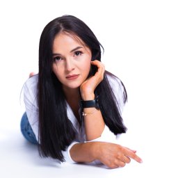 Oczami Zajawki Fotografia - Kobieta z ciemnymi włosami leży na białym tle, ubrana w białą koszulę i dżinsy. Na nadgarstku widoczny zegarek i bransoletka. Ujęcie z góry, skupione na twarzy.