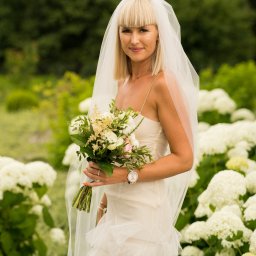 Oczami Zajawki Fotografia - Panna młoda w zwiewnej sukni, z welonem i bukietem, pozuje na tle kwitnących hortensji. Delikatne światło podkreśla naturalny makijaż i subtelną biżuterię.