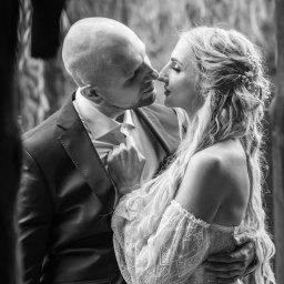 Oczami Zajawki Fotografia - Czarno-białe zbliżenie pary młodej w romantycznym uścisku. Pan młody w garniturze, panna młoda w koronkowej sukni z kwiatami we włosach. Delikatne światło podkreśla emocje.