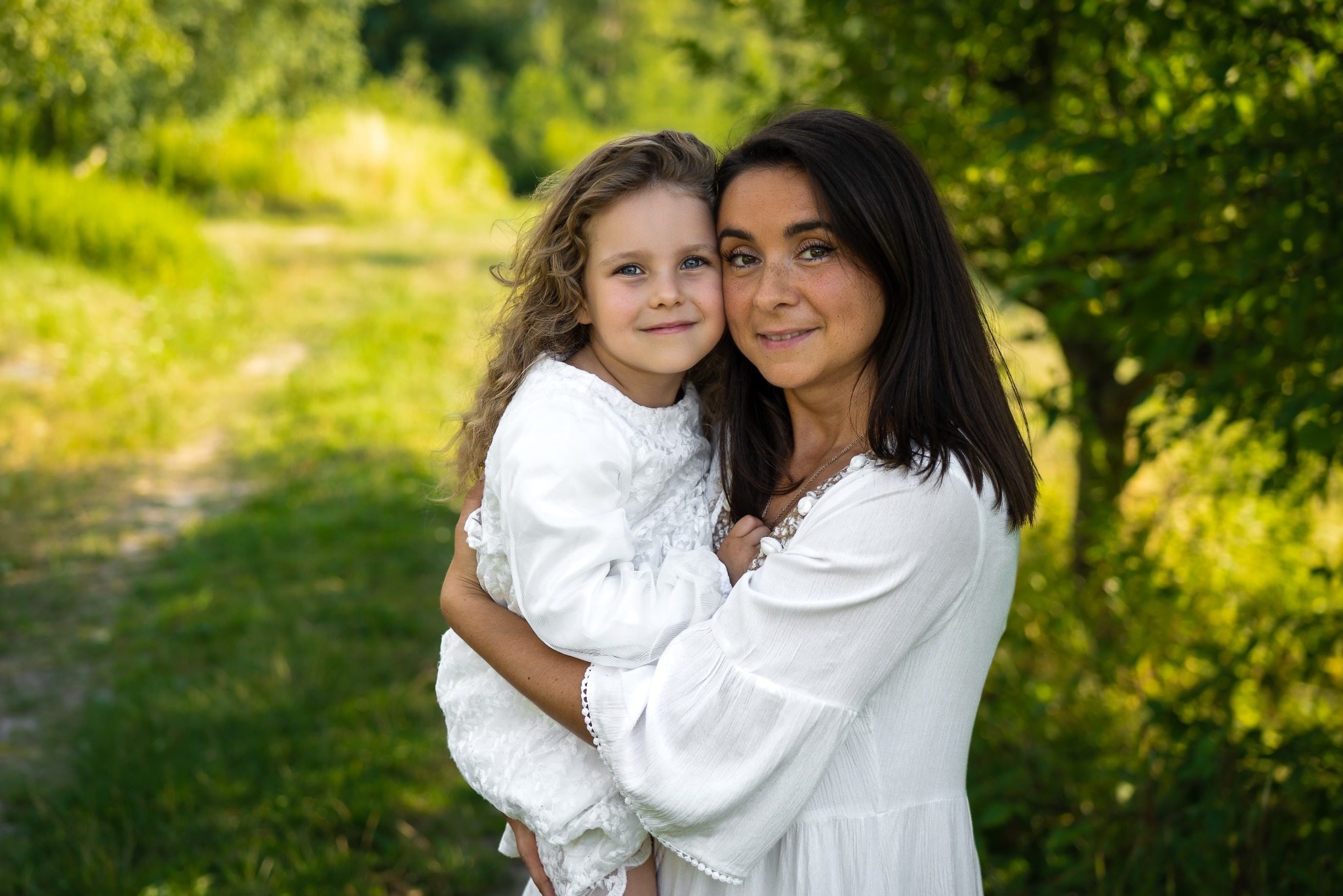 Portret matki z córką w białych sukienkach na tle zieleni. Uśmiechnięte twarze, naturalne światło, miękkie tło. Romantyczna atmosfera, bliskość i ciepło rodzinne.