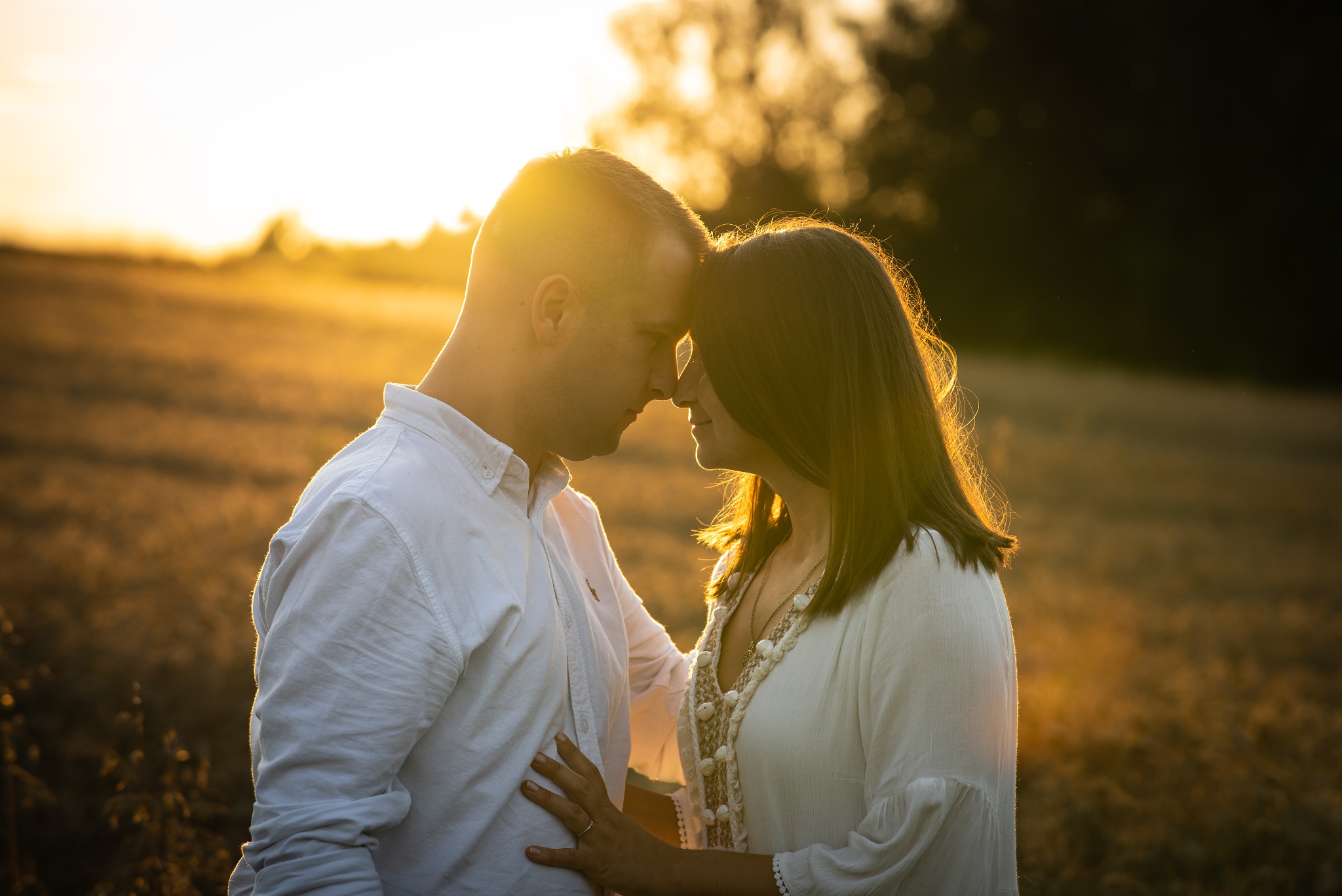 Para zakochanych, stykająca się czołami na tle zachodzącego słońca, w otoczeniu pola. Romantyczna atmosfera, ciepłe barwy, delikatne światło podkreśla intymność chwili.
