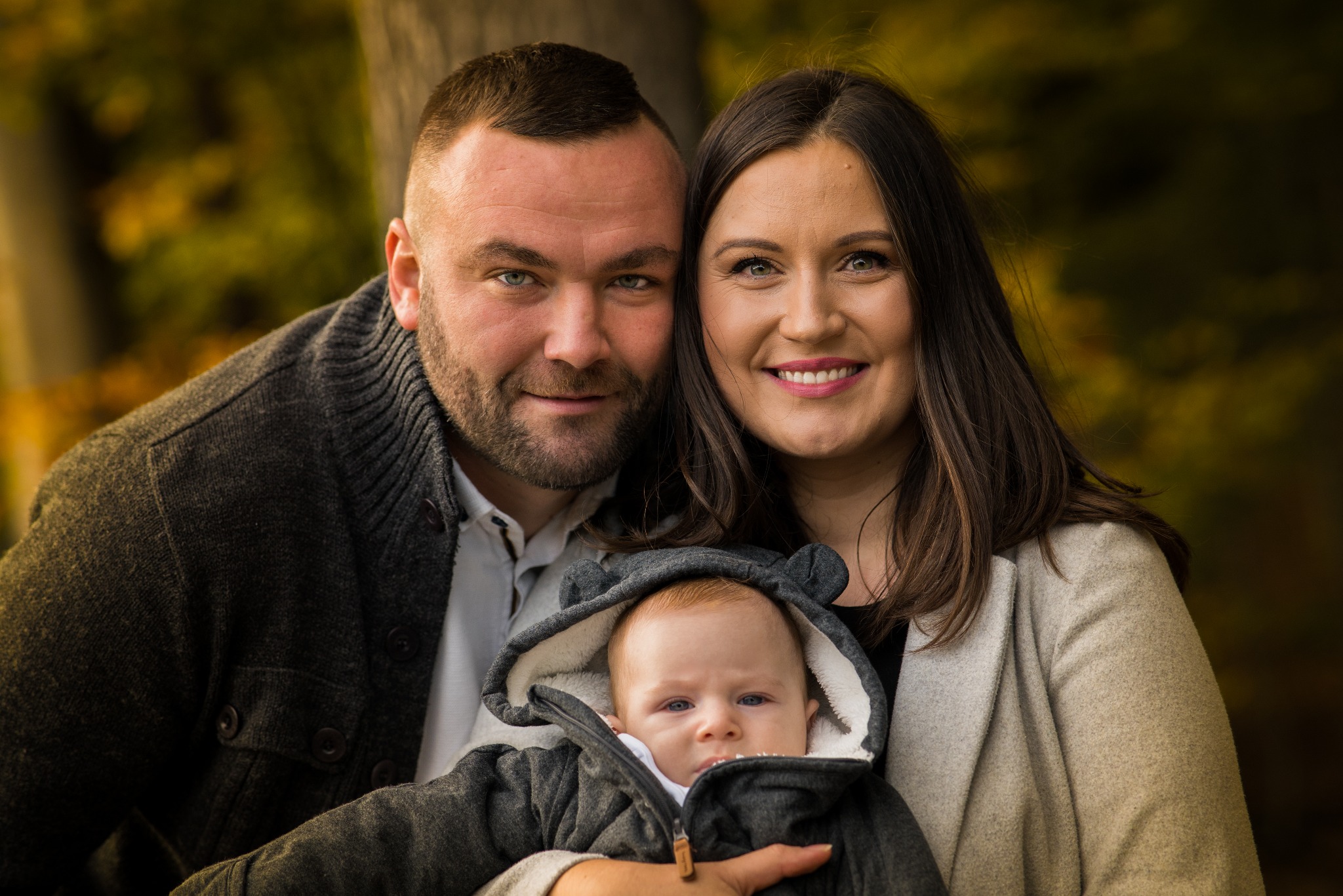 Portret uśmiechniętej rodziny: ojciec z brodą, matka z długimi włosami i niemowlę w kurtce z kapturem na tle jesiennych drzew.