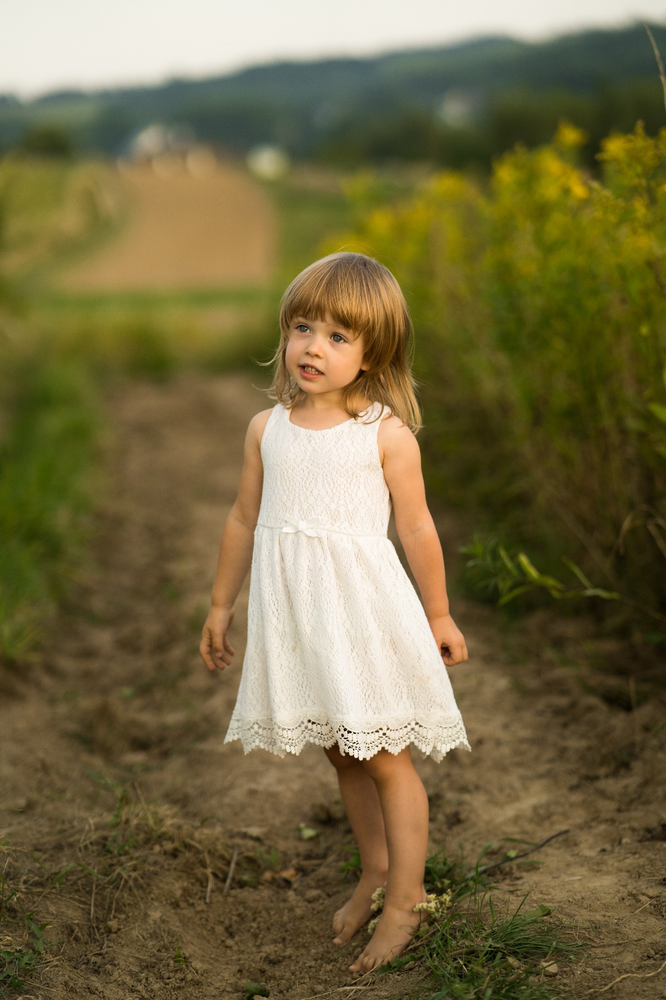 Urocza, bosa dziewczynka w białej sukience stoi na polnej drodze, spoglądając w górę. W tle zieleń i rozmyty krajobraz. Delikatne, naturalne światło.