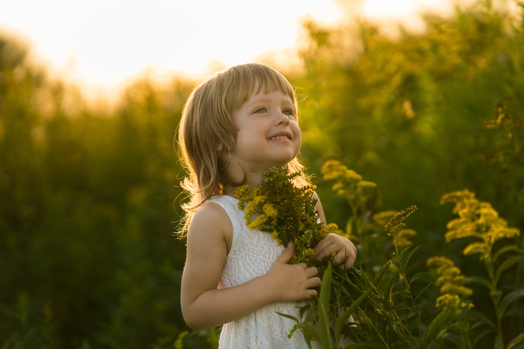 Uśmiechnięta dziewczynka w białej sukience trzyma bukiet żółtych kwiatów na tle zieleni w ciepłym, zachodzącym słońcu. Delikatne światło podkreśla naturalność sceny.