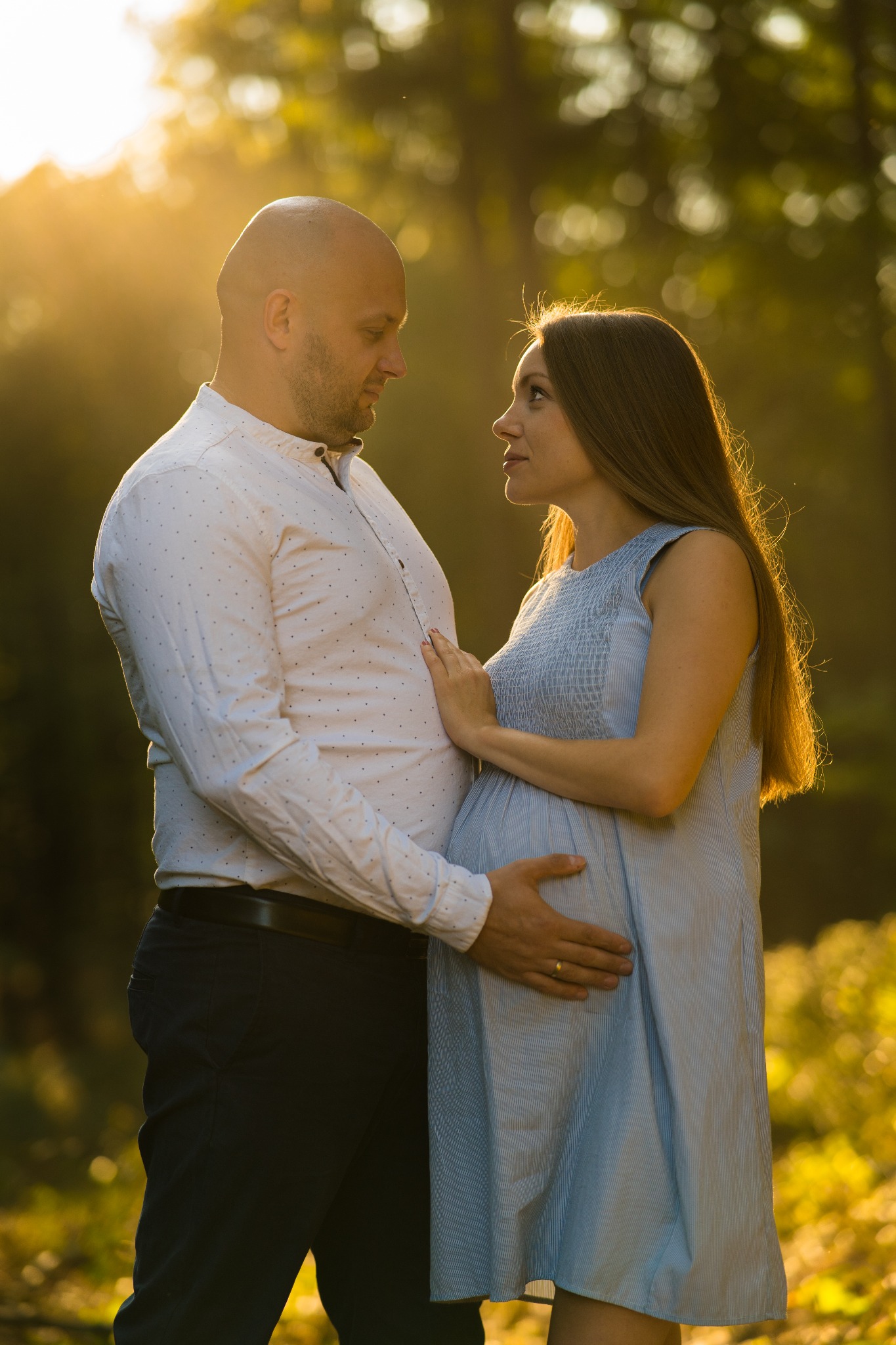 Zakochana para w promieniach zachodzącego słońca. Kobieta w jasnoniebieskiej sukience, mężczyzna w białej koszuli czule obejmujący ciążowy brzuszek. Romantyczna atmosfera.