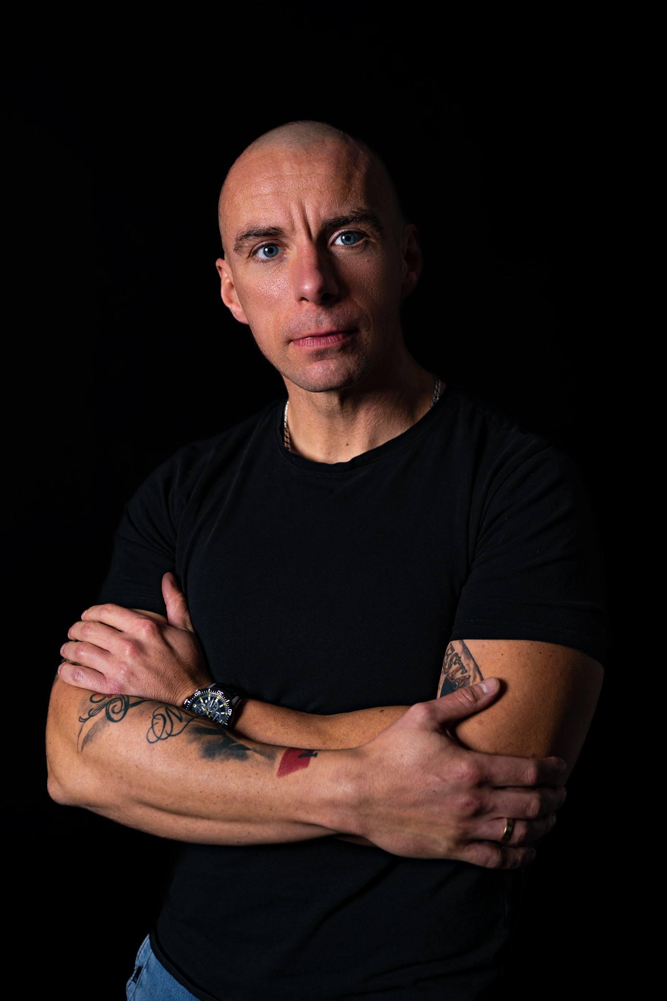 Portret mężczyzny z niebieskimi oczami na czarnym tle. Widoczne tatuaże na ramionach, zegarek i obrączka. Ubrany w czarny t-shirt, z założonymi rękami.