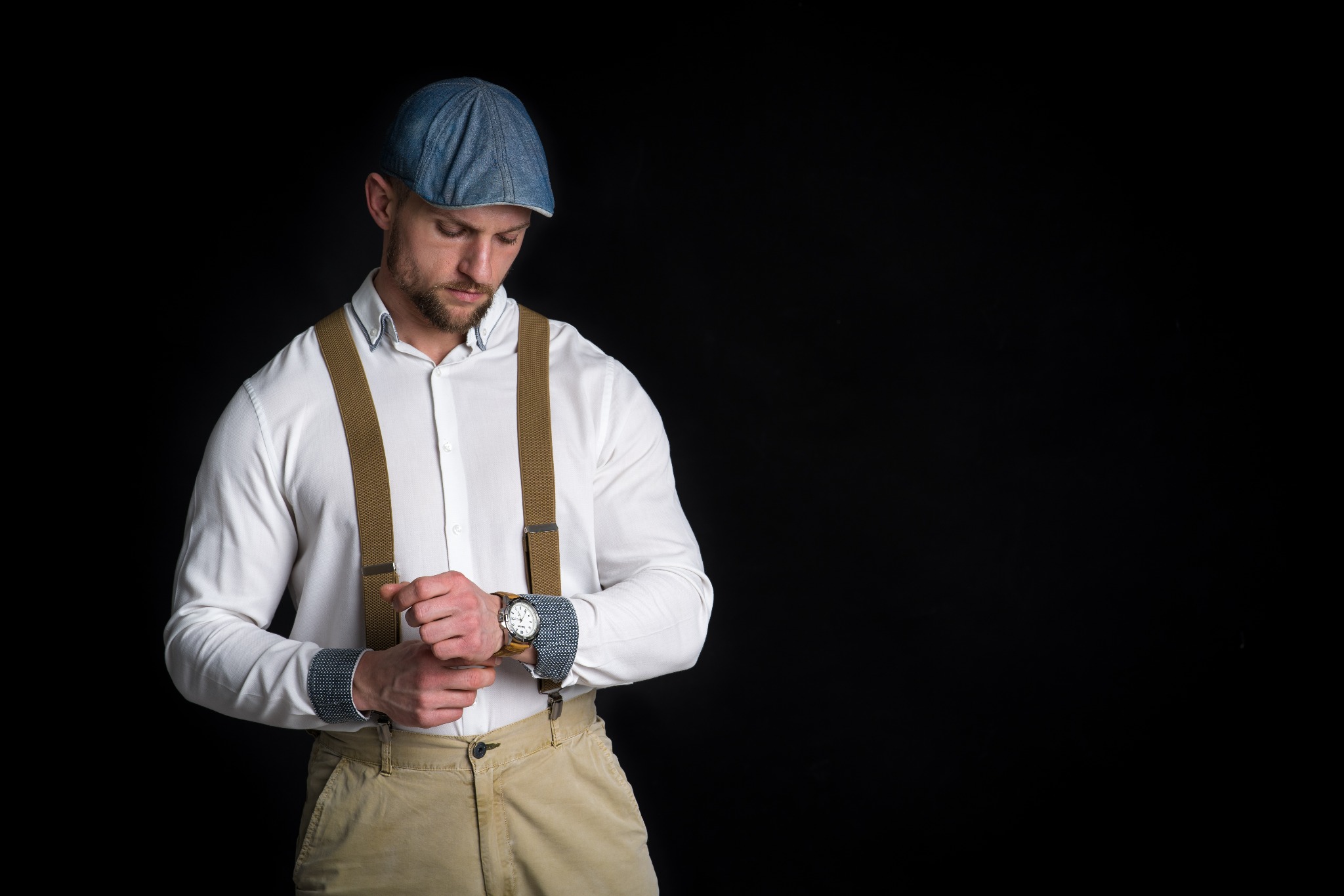 Mężczyzna w kaszkiecie i szelkach, z brodą, spogląda na zegarek na nadgarstku. Biała koszula z podwiniętymi rękawami, beżowe spodnie. Czarne tło, studyjne oświetlenie.