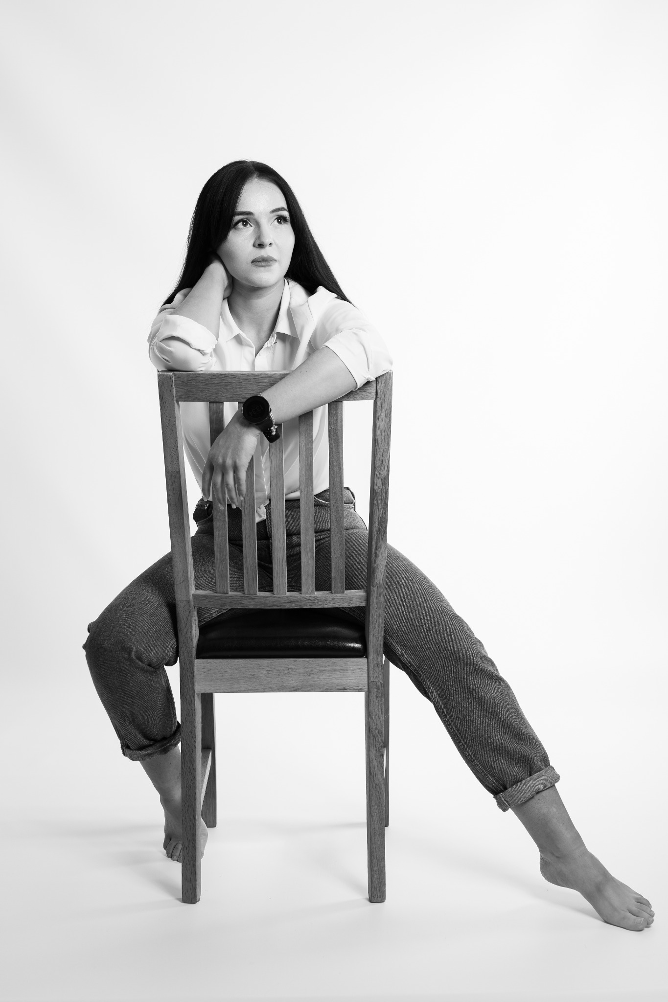 Czarno-białe zdjęcie kobiety siedzącej na krześle, z jedną nogą wyciągniętą, w koszuli i dżinsach, zamyślona poza na jasnym tle. Widoczny zegarek na ręce.