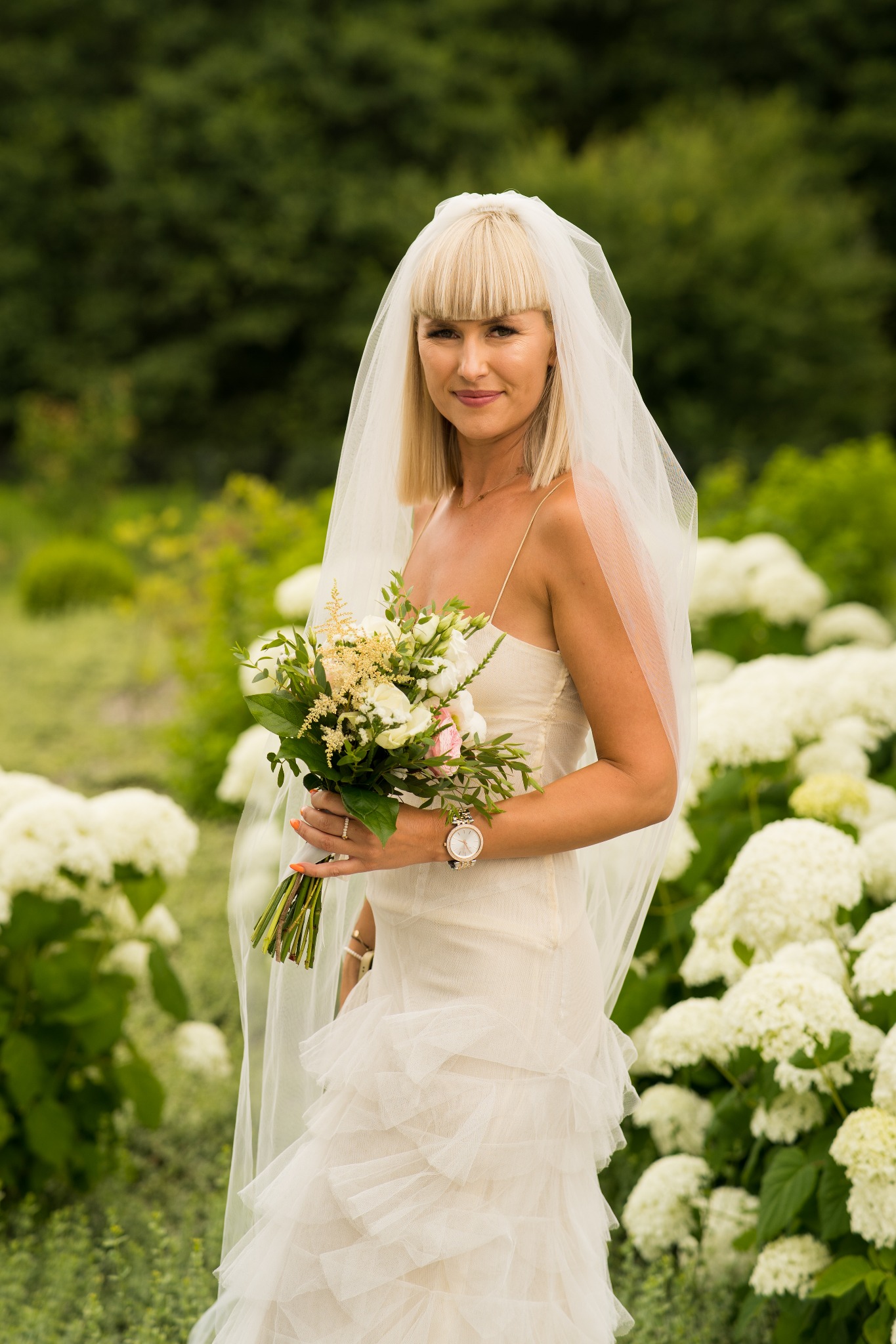Panna młoda w zwiewnej sukni, z welonem i bukietem, pozuje na tle kwitnących hortensji. Delikatne światło podkreśla naturalny makijaż i subtelną biżuterię.