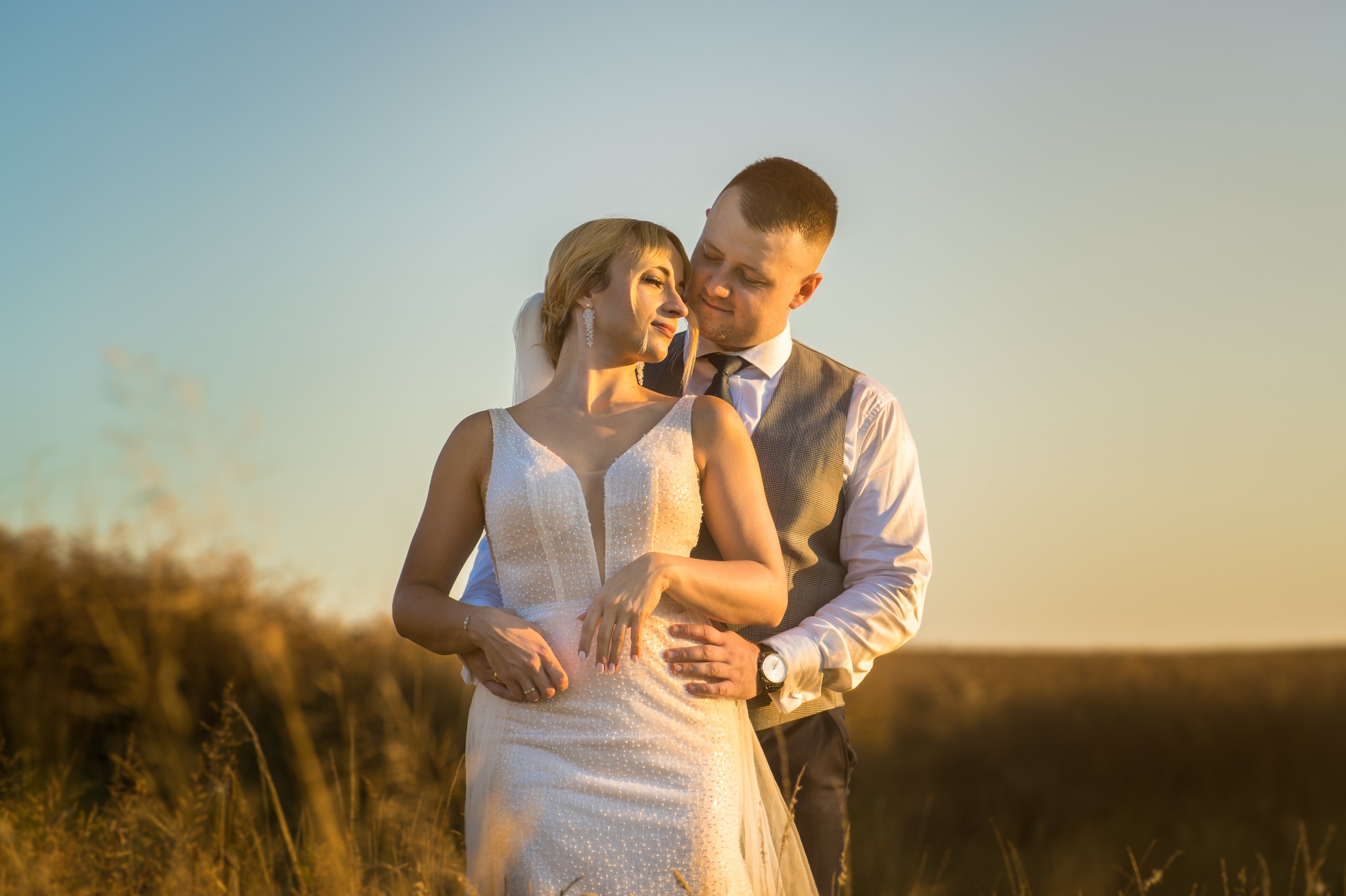 Para młoda w objęciach na tle łąki w ciepłym świetle zachodzącego słońca. Panna młoda w sukni z głębokim dekoltem, pan młody w kamizelce.