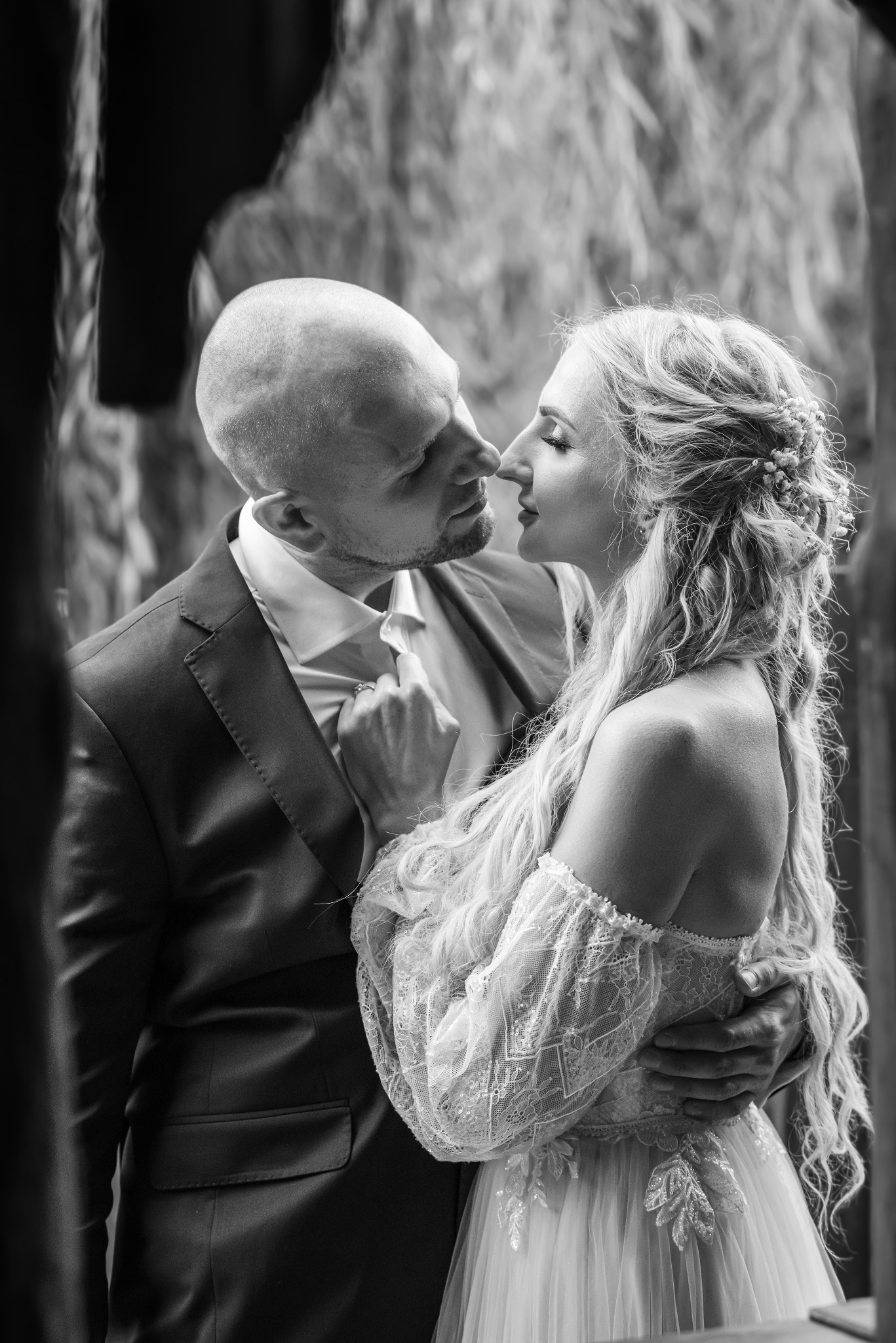 Czarno-białe zbliżenie pary młodej w romantycznym uścisku. Pan młody w garniturze, panna młoda w koronkowej sukni z kwiatami we włosach. Delikatne światło podkreśla emocje.