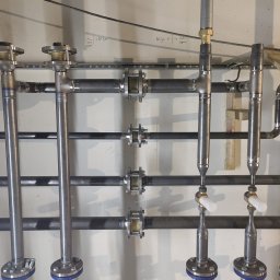 Euro Instal - Instalacja przemysłowa z rur metalowych, zaworów z niebieskimi pokrętłami i złączek, zamontowana na jasnej ścianie z widocznymi napisami.