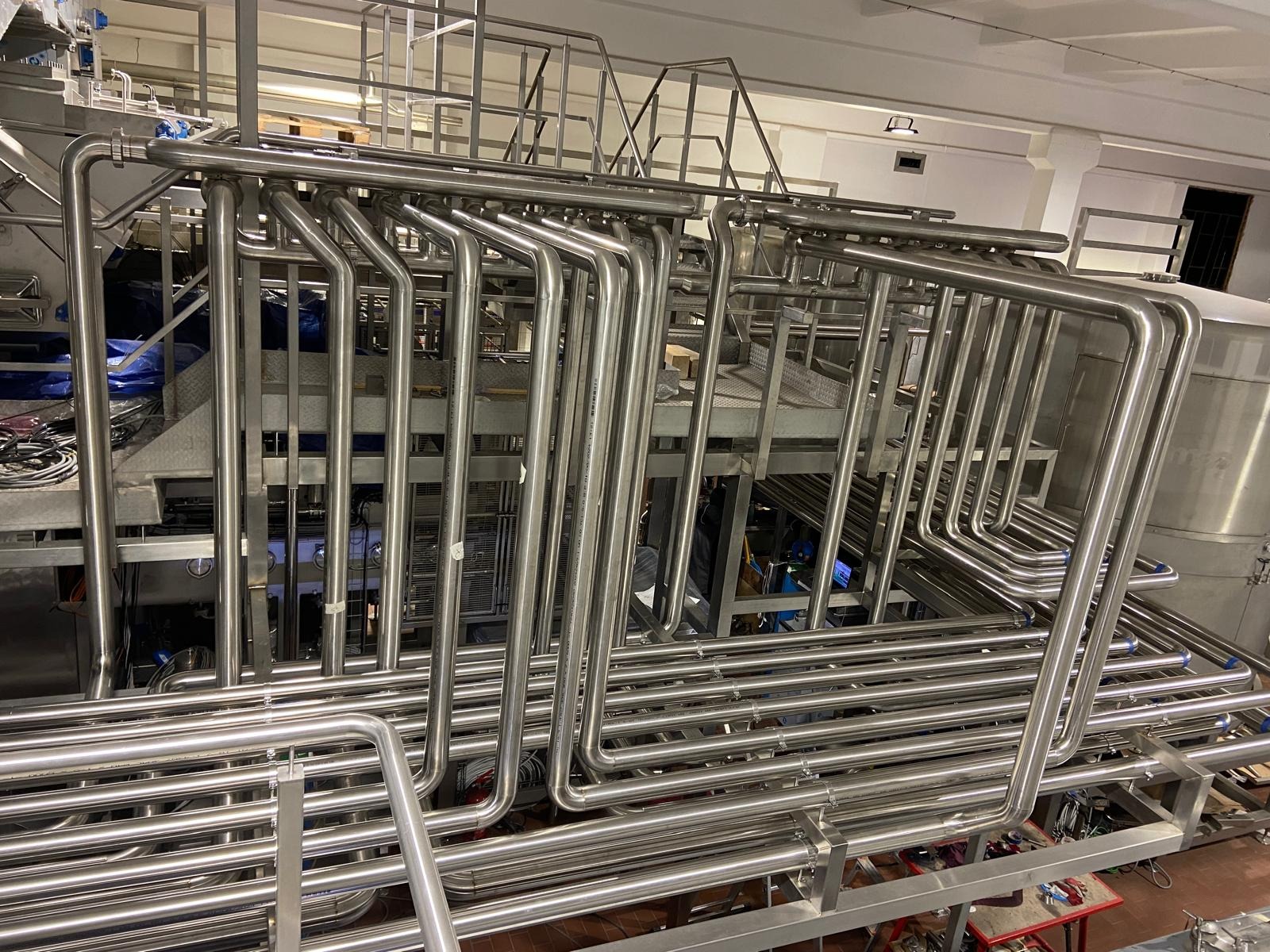 Kompleksowa instalacja rur ze stali nierdzewnej w hali przemysłowej, widoczne liczne złącza i podesty, oświetlenie górne