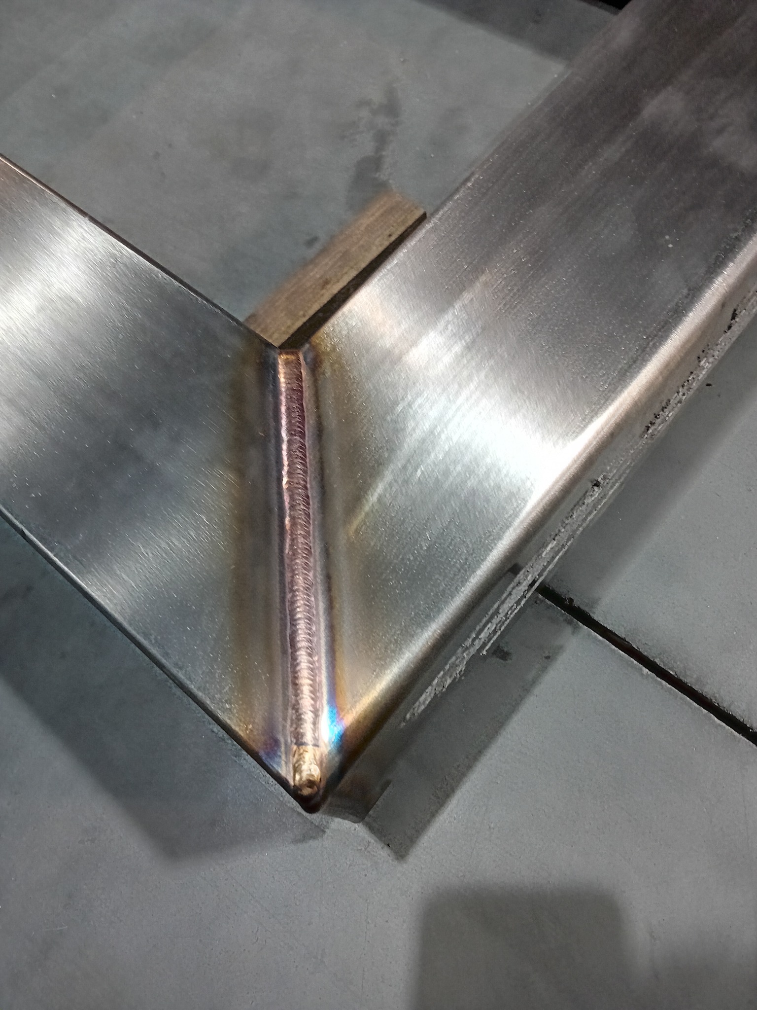 Spoina na kątowniku ze stali nierdzewnej, widoczne przebarwienia termiczne, wsparta na drewnianym klocku.