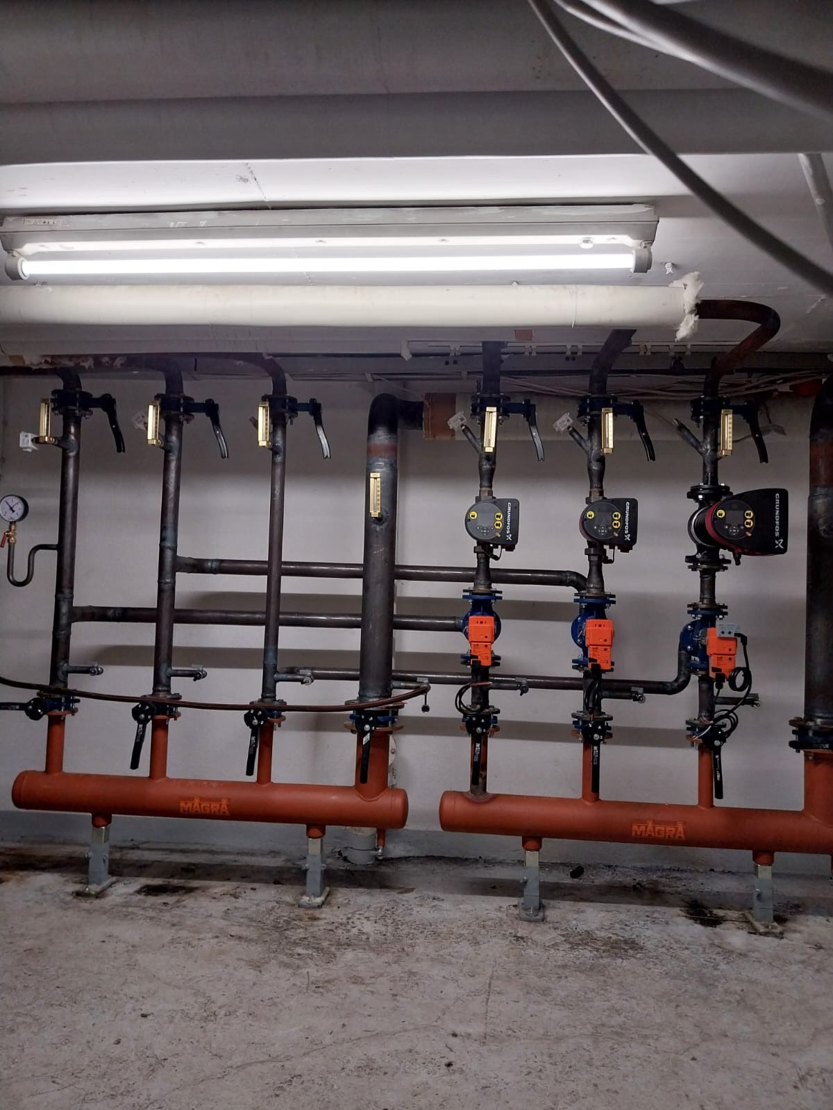 Instalacja centralnego ogrzewania z rurami, pompami obiegowymi Grundfos i zaworami, umieszczona w piwnicy pod oświetleniem jarzeniowym.