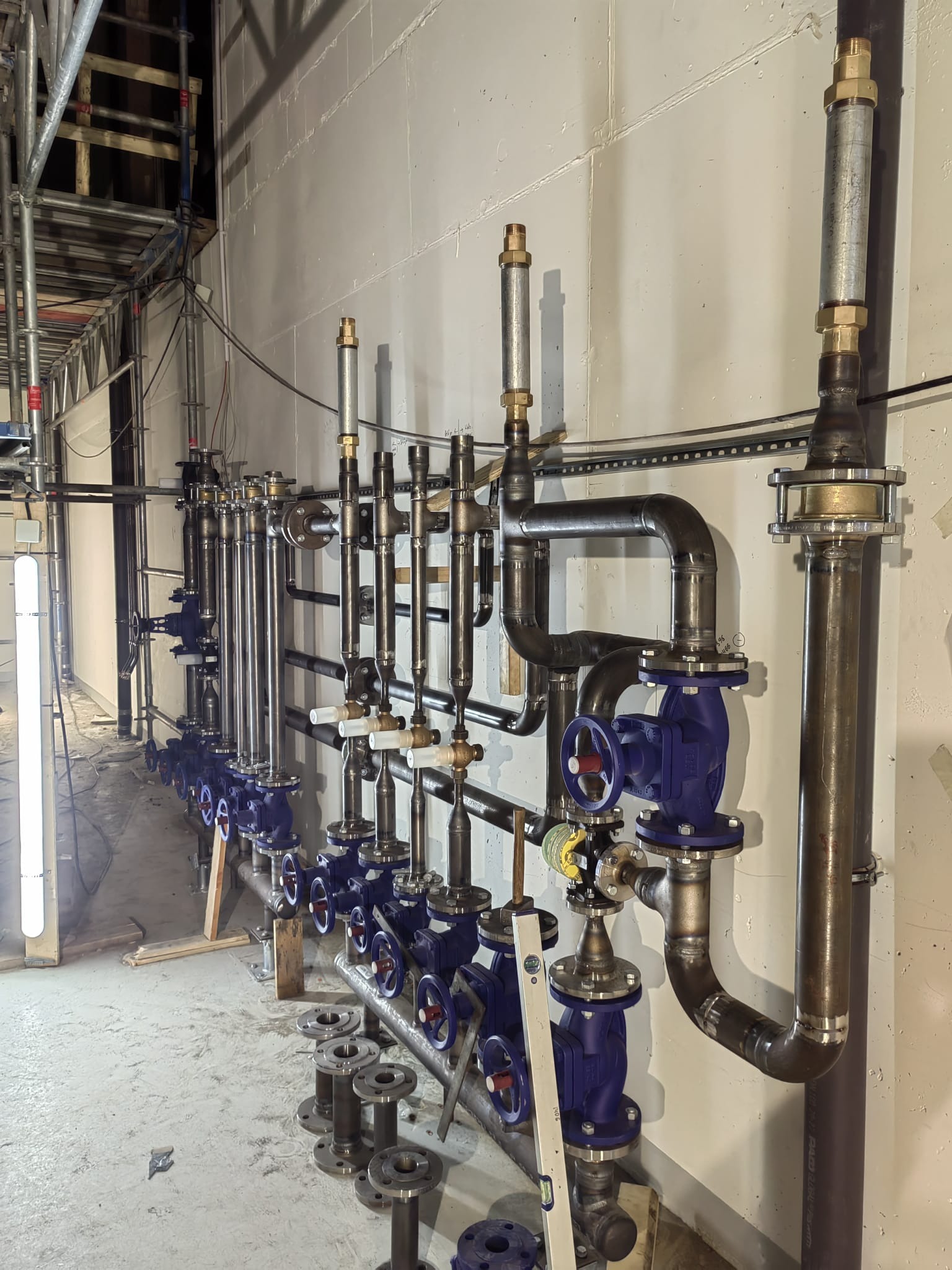 Szereg metalowych rur z niebieskimi zaworami i spawami na ścianie, widoczne rusztowanie w tle, poziomica oparta o rury, przygotowane elementy do montażu na podłodze.