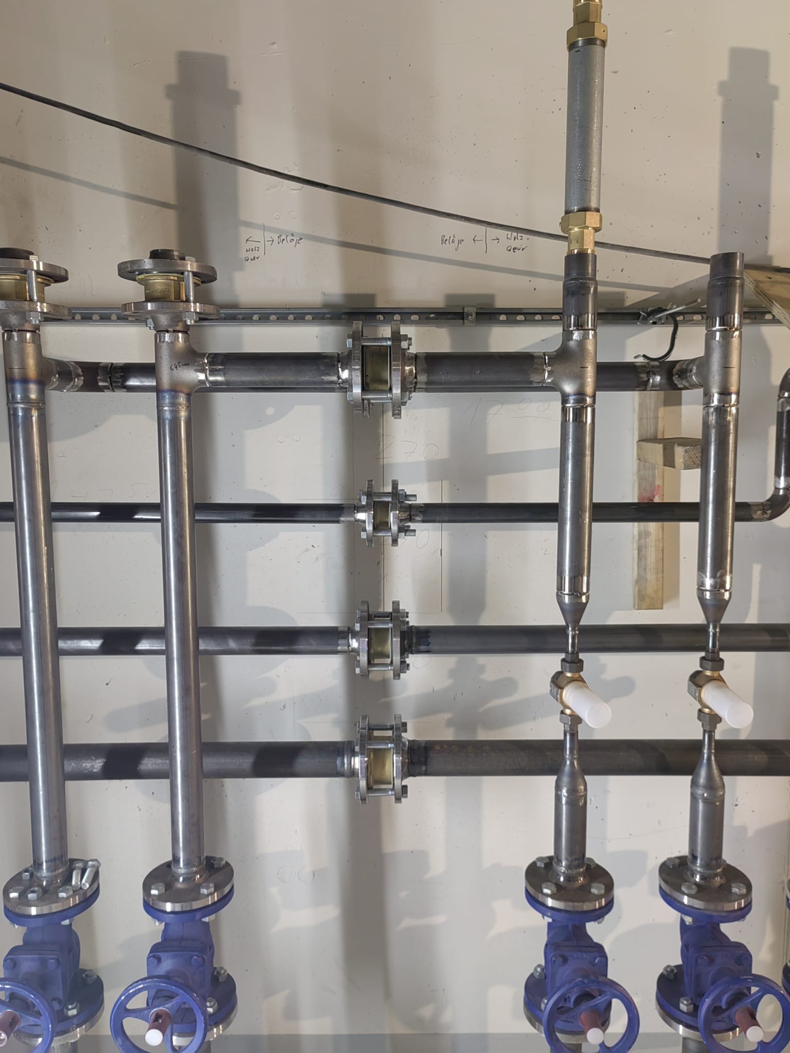 Instalacja przemysłowa z rur metalowych, zaworów z niebieskimi pokrętłami i złączek, zamontowana na jasnej ścianie z widocznymi napisami.