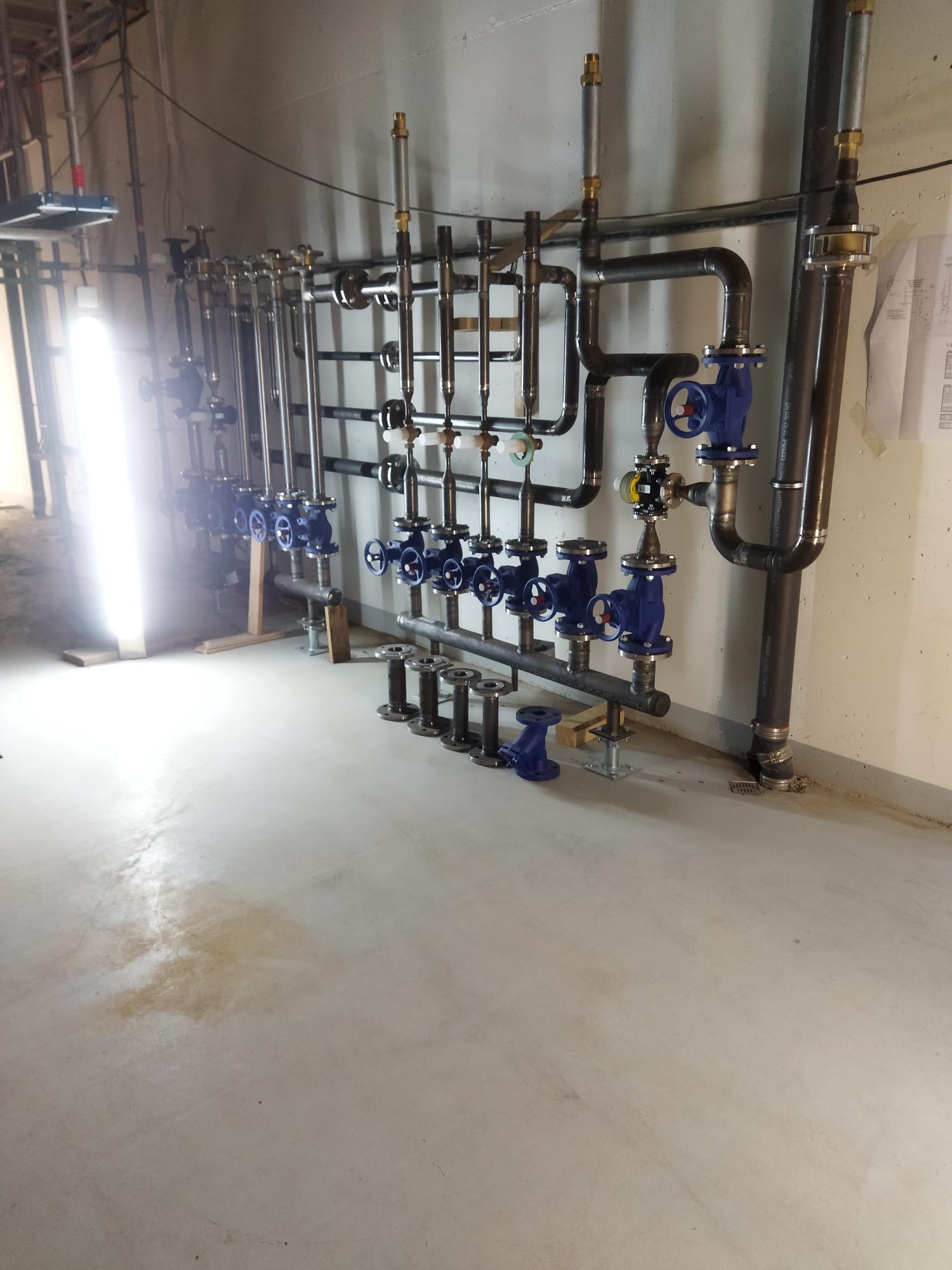 Instalacja przemysłowa: Rząd rur z niebieskimi zaworami, metalowe wsporniki i spawane połączenia na tle betonowej ściany i posadzki.