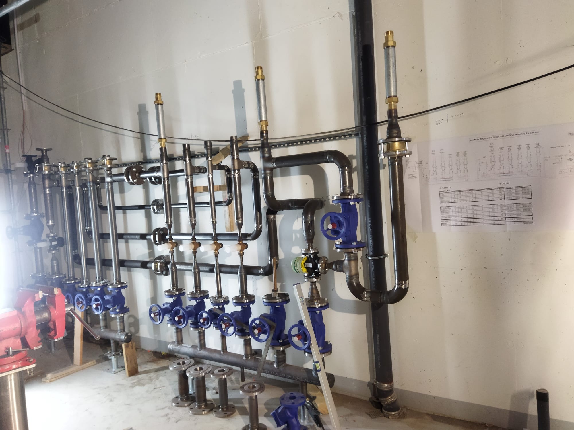 Instalacja przemysłowa z rur i zaworów w piwnicy, widoczne elementy montażowe, plan instalacji na ścianie i narzędzie do obróbki rur