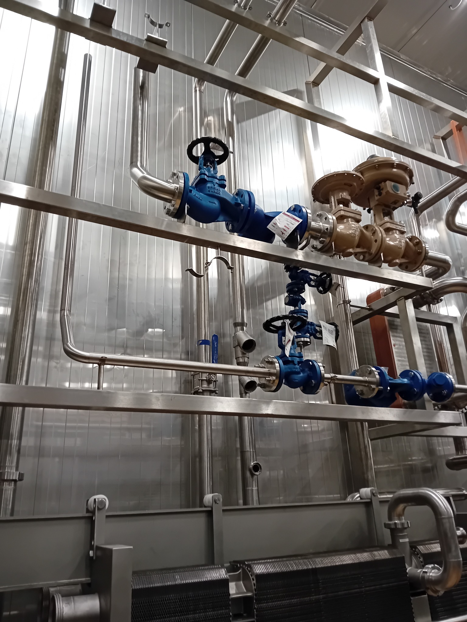 Instalacja przemysłowa z rurami i zaworami w odcieniach niebieskiego i brązu, zamontowana na metalowej konstrukcji wsporczej na tle blaszanej ściany.