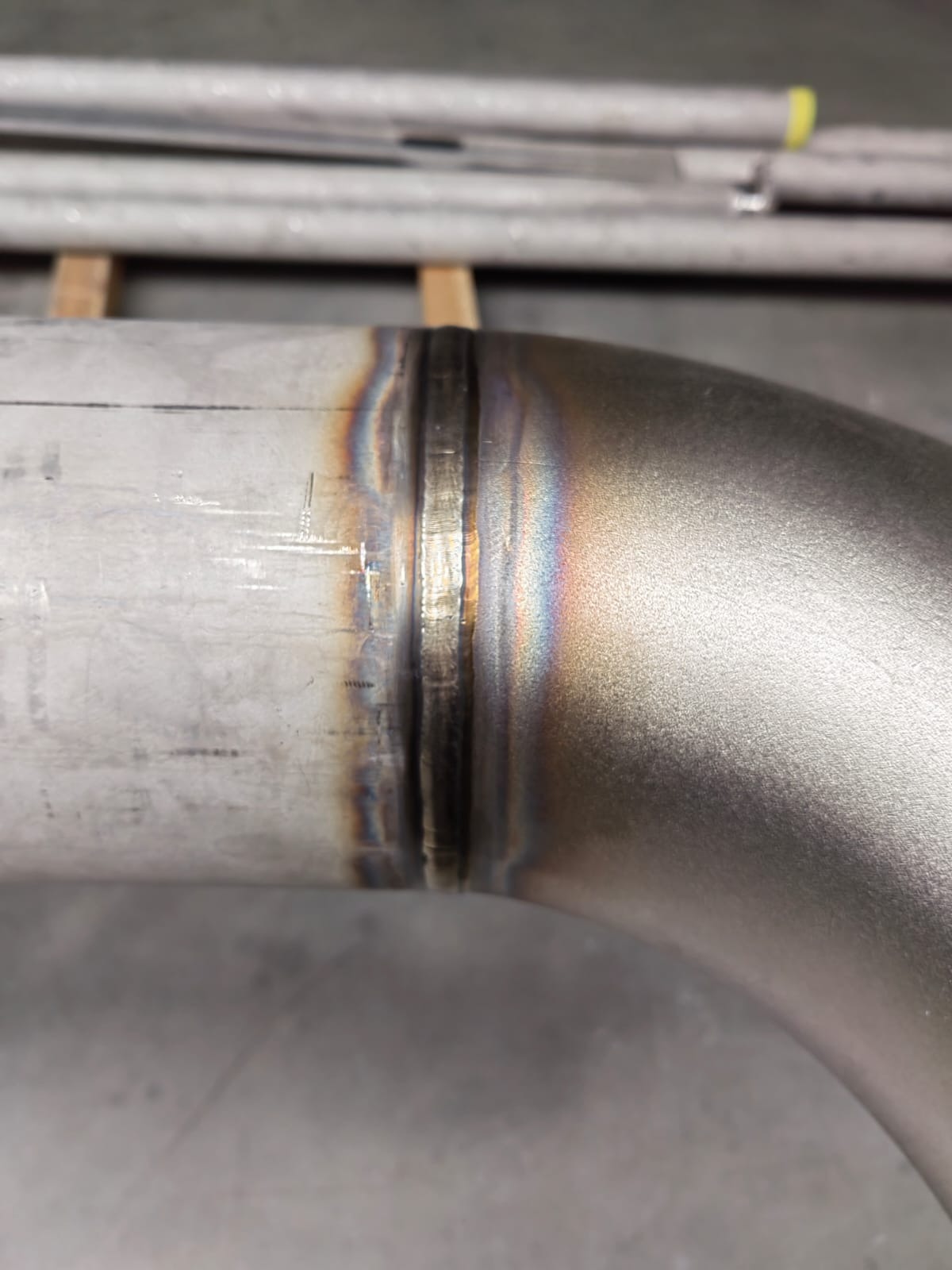 Szczegółowe zbliżenie na spawany, nierdzewny kolanko rury, z widocznymi śladami obróbki termicznej i tęczowymi przebarwieniami w strefie spawu.