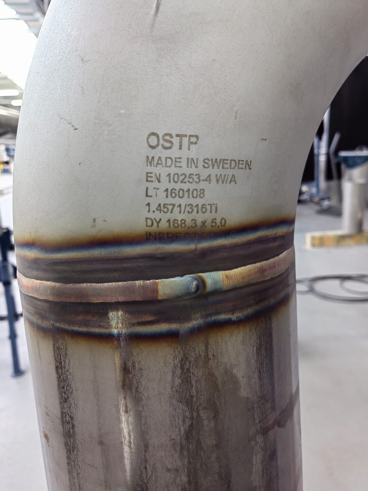 Spoina na rurze ze stali nierdzewnej z widocznym napisem 'OSTP MADE IN SWEDEN' i specyfikacją techniczną, ukazująca charakterystyczne przebarwienia po procesie łączenia.