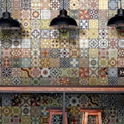 Wielobarwna ściana z płytek o zróżnicowanych wzorach, oświetlona trzema czarnymi lampami wiszącymi z metalowymi kloszami, uzupełniona drewnianym blatem i dwoma stołkami barowymi.