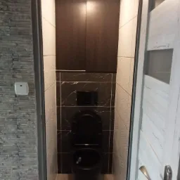 Wąskie ujęcie wyremontowanej toalety z czarną armaturą i marmurkowymi płytkami, drewnianą szafką nad toaletą i dekoracyjną ścianą z kamienia po lewej stronie.