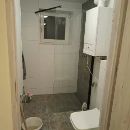 Wnętrze łazienki w trakcie prac: widoczny biały bojler na szarej ścianie z płytek, biała toaleta, wiadro z wodą i mop oraz butelka płynu do naczyń.