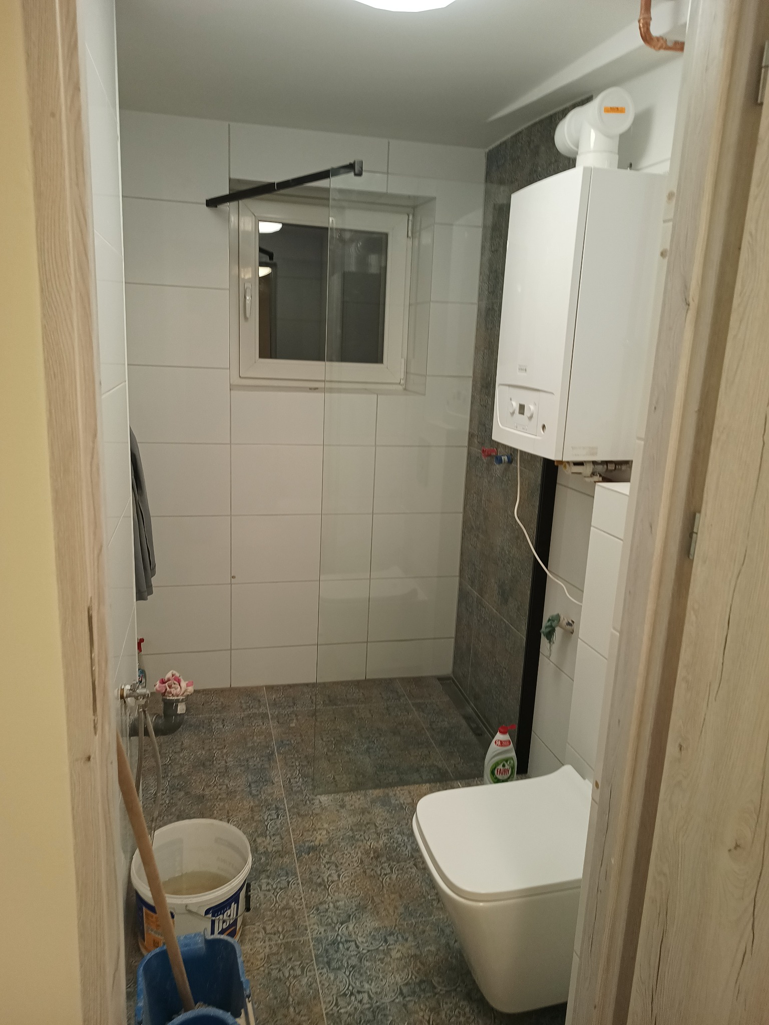 Wnętrze łazienki w trakcie prac: widoczny biały bojler na szarej ścianie z płytek, biała toaleta, wiadro z wodą i mop oraz butelka płynu do naczyń.