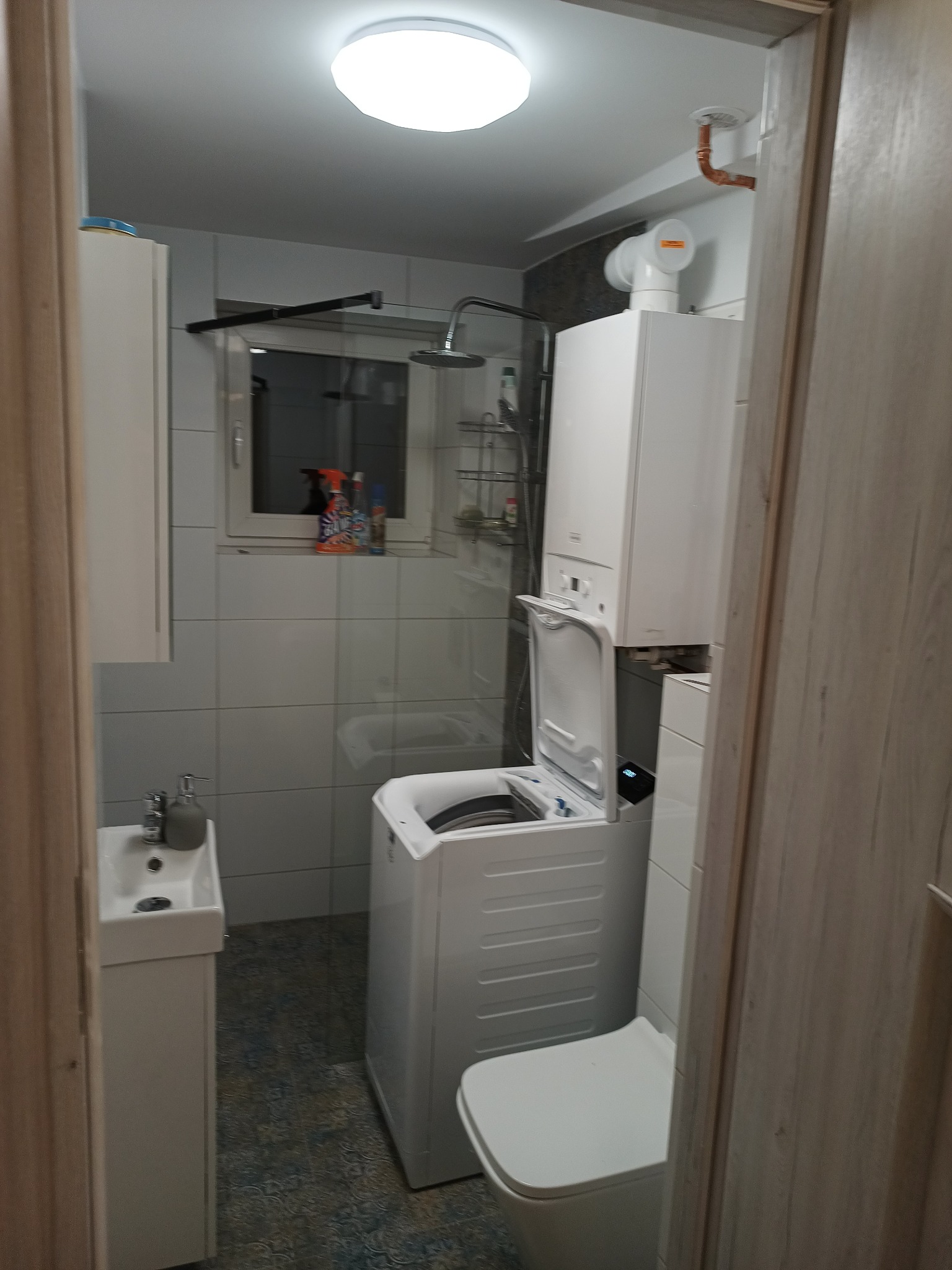 Mała łazienka z białą umywalką na szafce, pralką z otwartymi drzwiczkami, toaletą, bojlerem gazowym i kabiną prysznicową w tle, oświetlona okrągłą lampą sufitową.