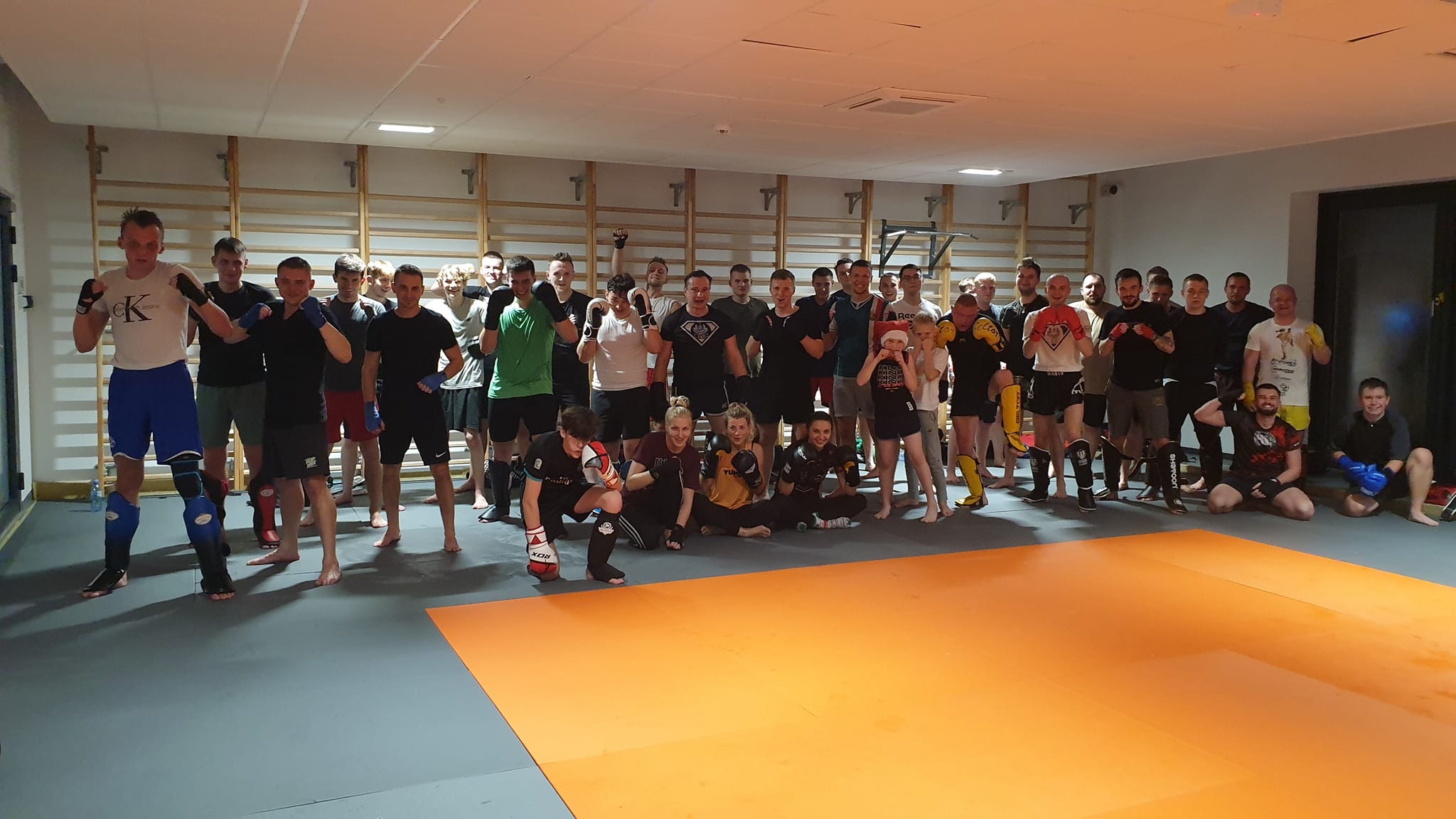 Grupa osób w różnym wieku, w strojach sportowych, pozuje do zdjęcia w sali z drabinkami gimnastycznymi i pomarańczową matą treningową.