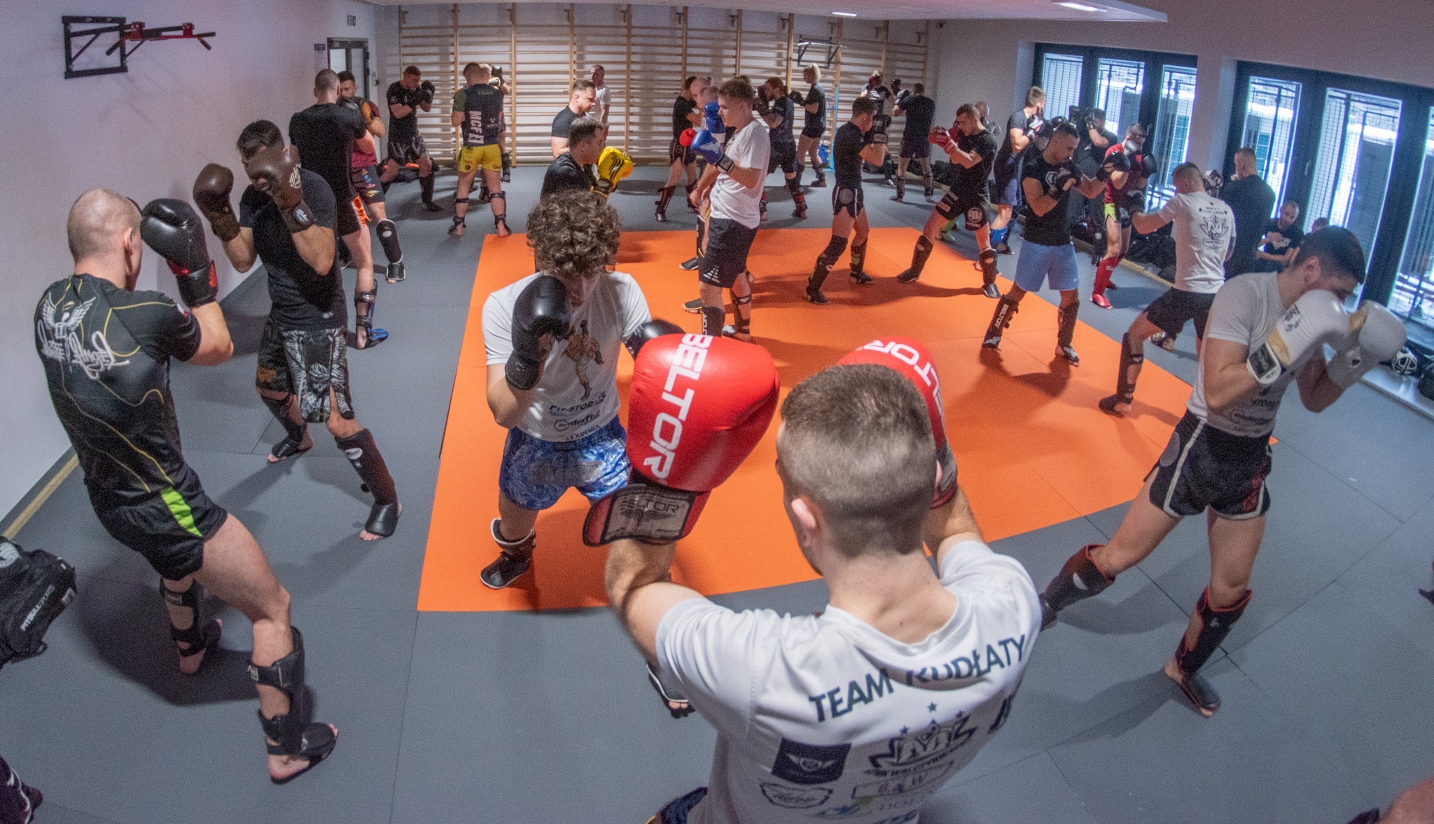 Sala treningowa z grupą mężczyzn w różnym sprzęcie ochronnym, trenujących sporty walki, widok z góry na pomarańczową matę i szare podłoże, w tle okna i ściany z drabinkami.