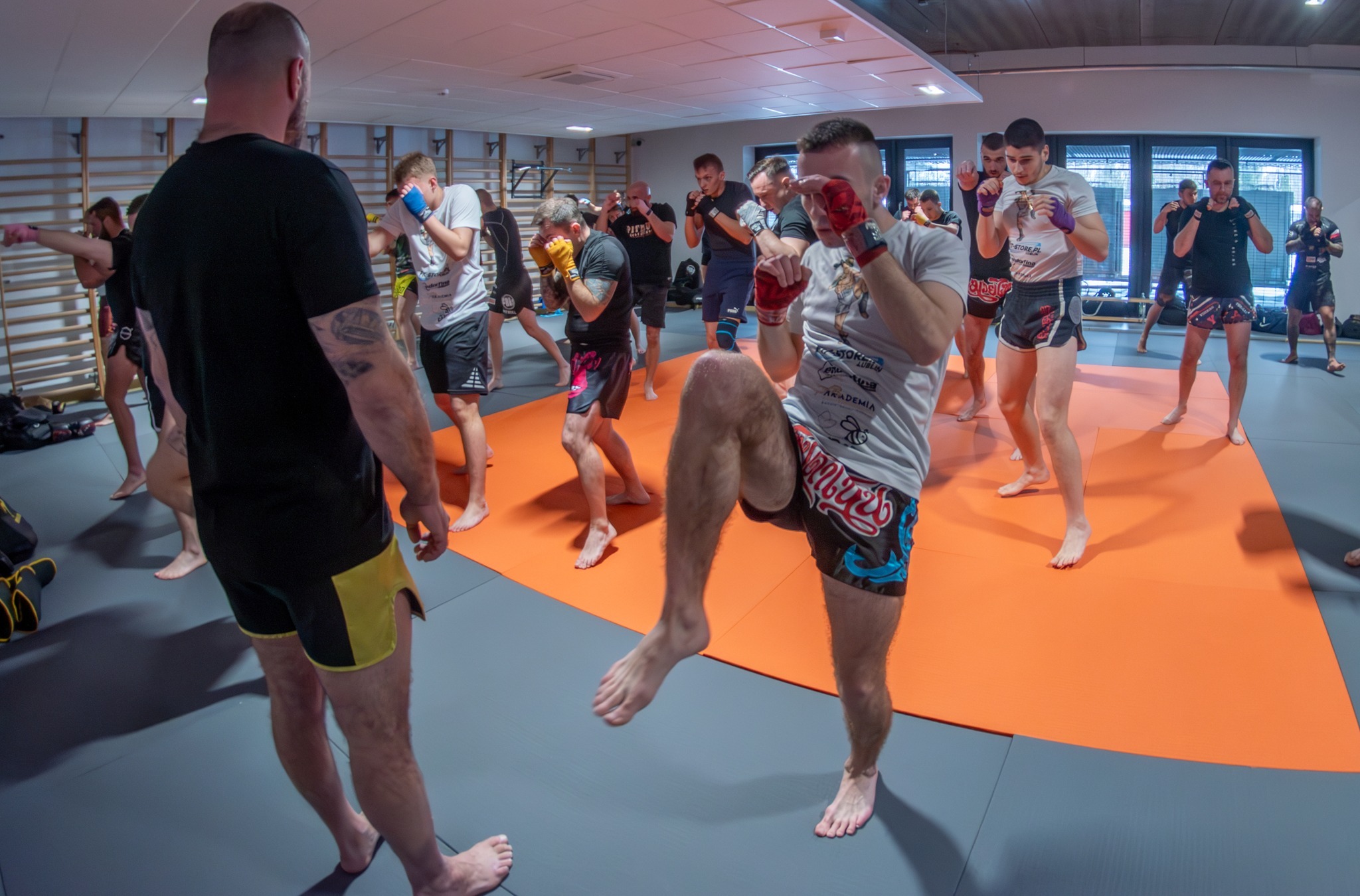 Grupa osób w sali ćwiczy kopnięcia kolanem, ubrani w spodenki i rękawice bokserskie, na pomarańczowej macie, z widocznymi drabinkami gimnastycznymi w tle.