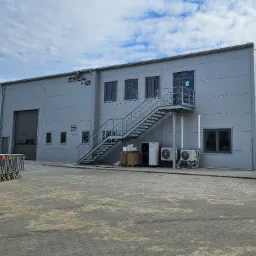 Szara hala przemysłowa z płyt warstwowych z zewnętrznymi schodami stalowymi, klimatyzatorami i bramą segmentową, widziana z zewnątrz na tle pochmurnego nieba.