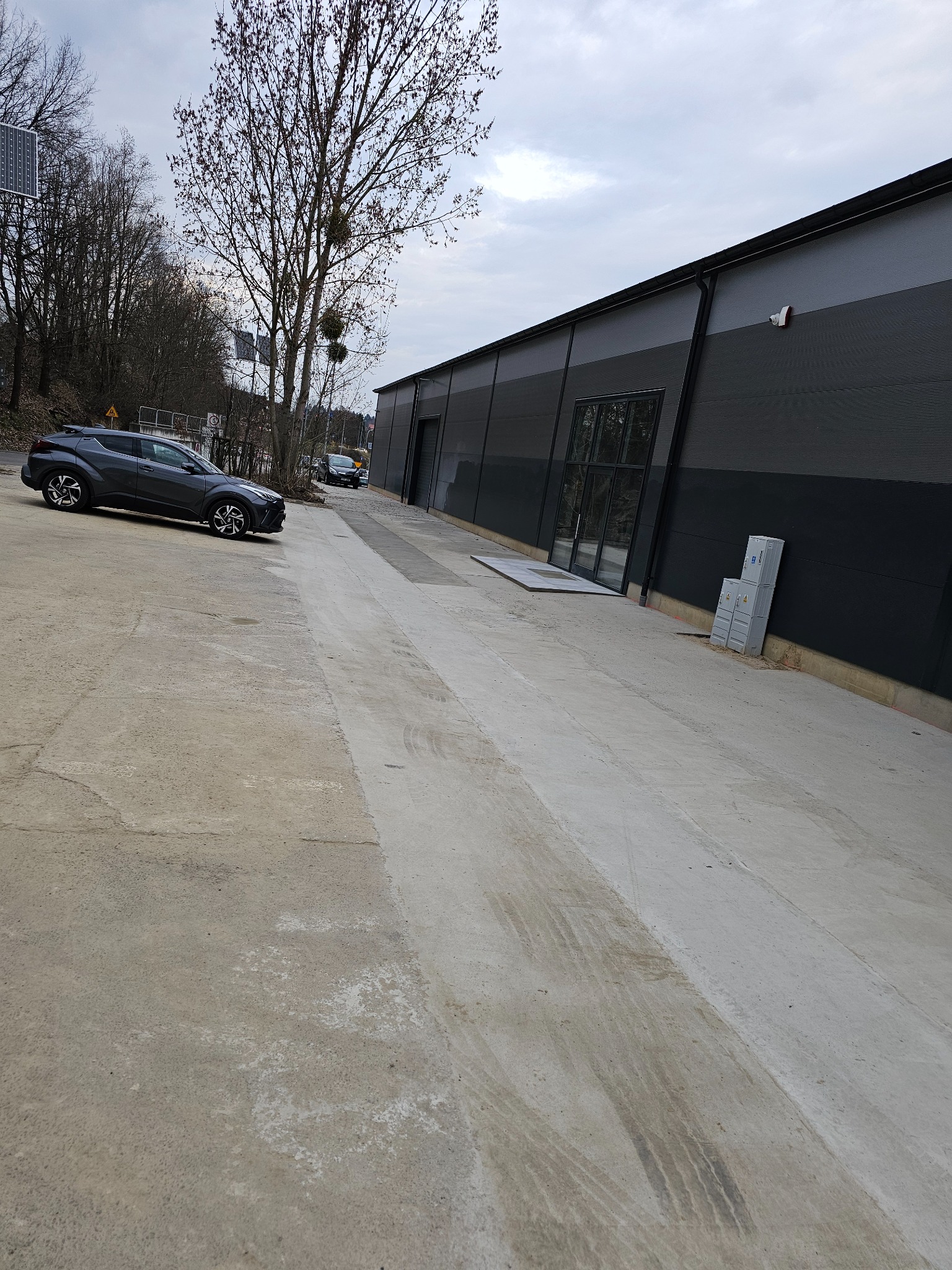 Zewnętrzny widok nowo wybudowanej hali z płyt warstwowych w szarej kolorystyce, z widocznymi drzwiami wejściowymi i zaparkowanym samochodem osobowym przed budynkiem.