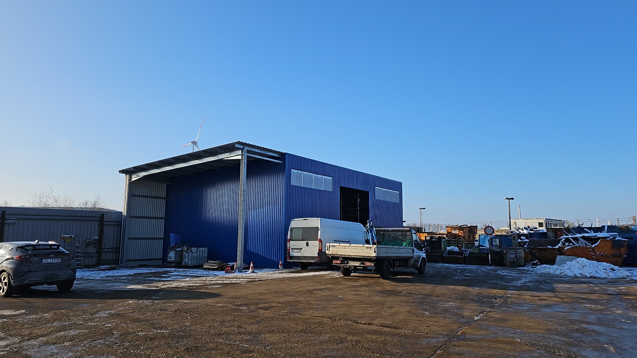 Niebieska hala z płyt warstwowych z otwartym wjazdem, zaparkowane busy, w tle turbina wiatrowa i kontenery.
