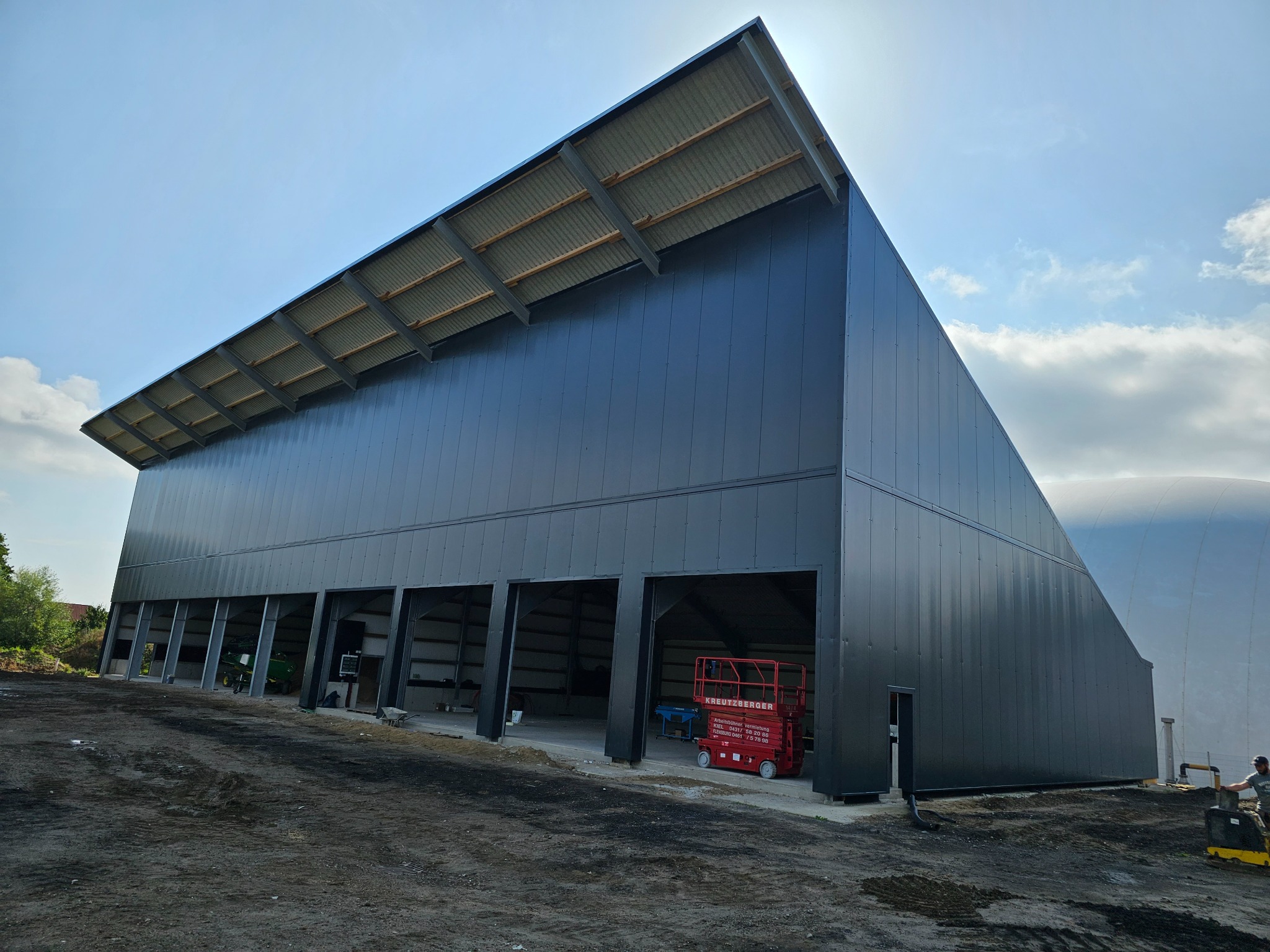 Nowoczesna hala stalowa z czarną elewacją z płyt warstwowych, widoczny podnośnik koszowy i maszyna budowlana, ziemisty teren.