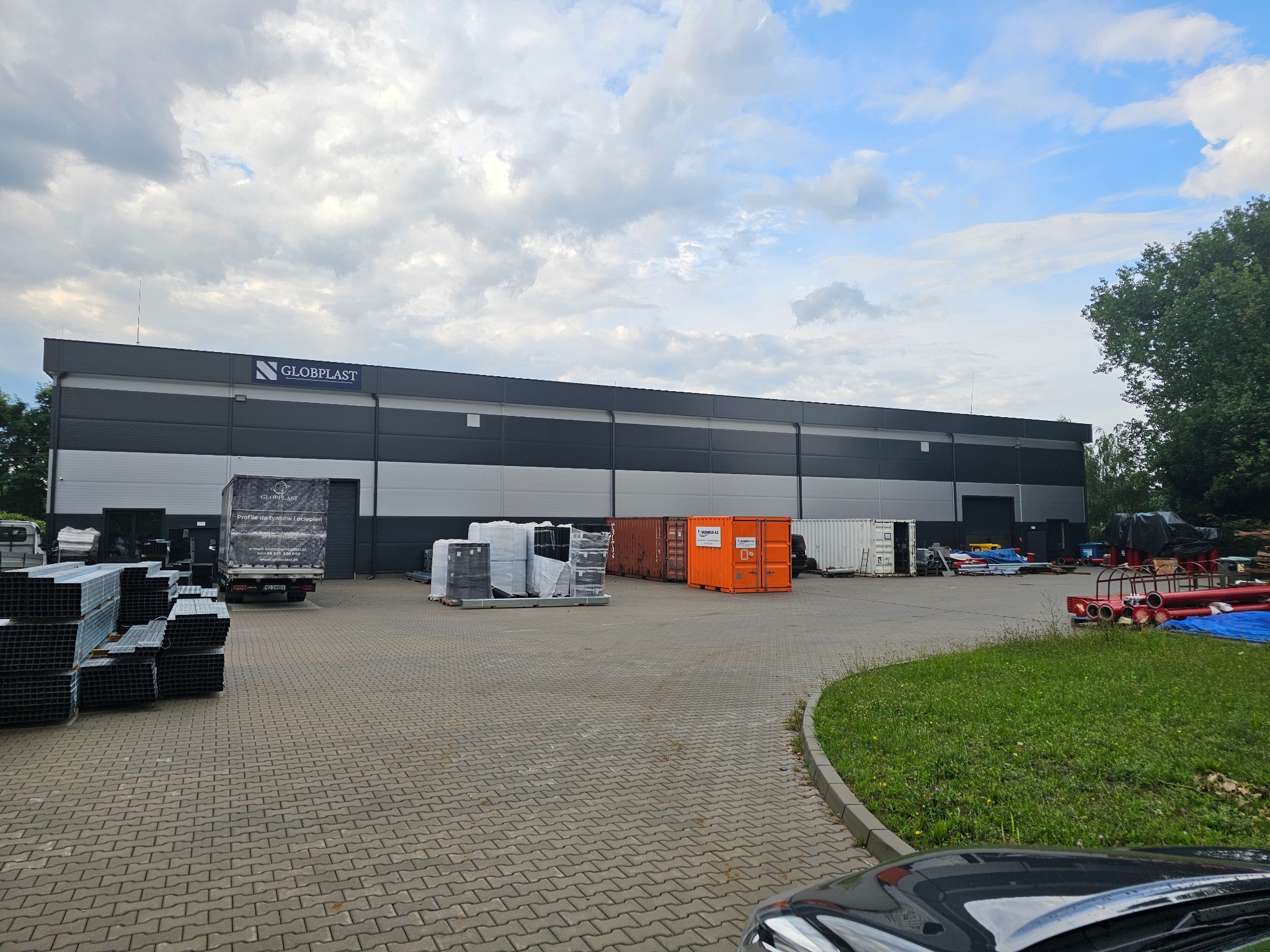 Gotowa hala przemysłowa z szarymi panelami i logo firmy Globplast, widoczny zaparkowany samochód dostawczy z reklamą firmy, składowane profile stalowe, kontenery magazynowe i inne materiały...