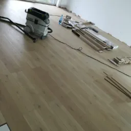 Układanie jasnych paneli podłogowych w pomieszczeniu, widoczny odkurzacz przemysłowy Festool, listwy przypodłogowe i opakowania paneli.