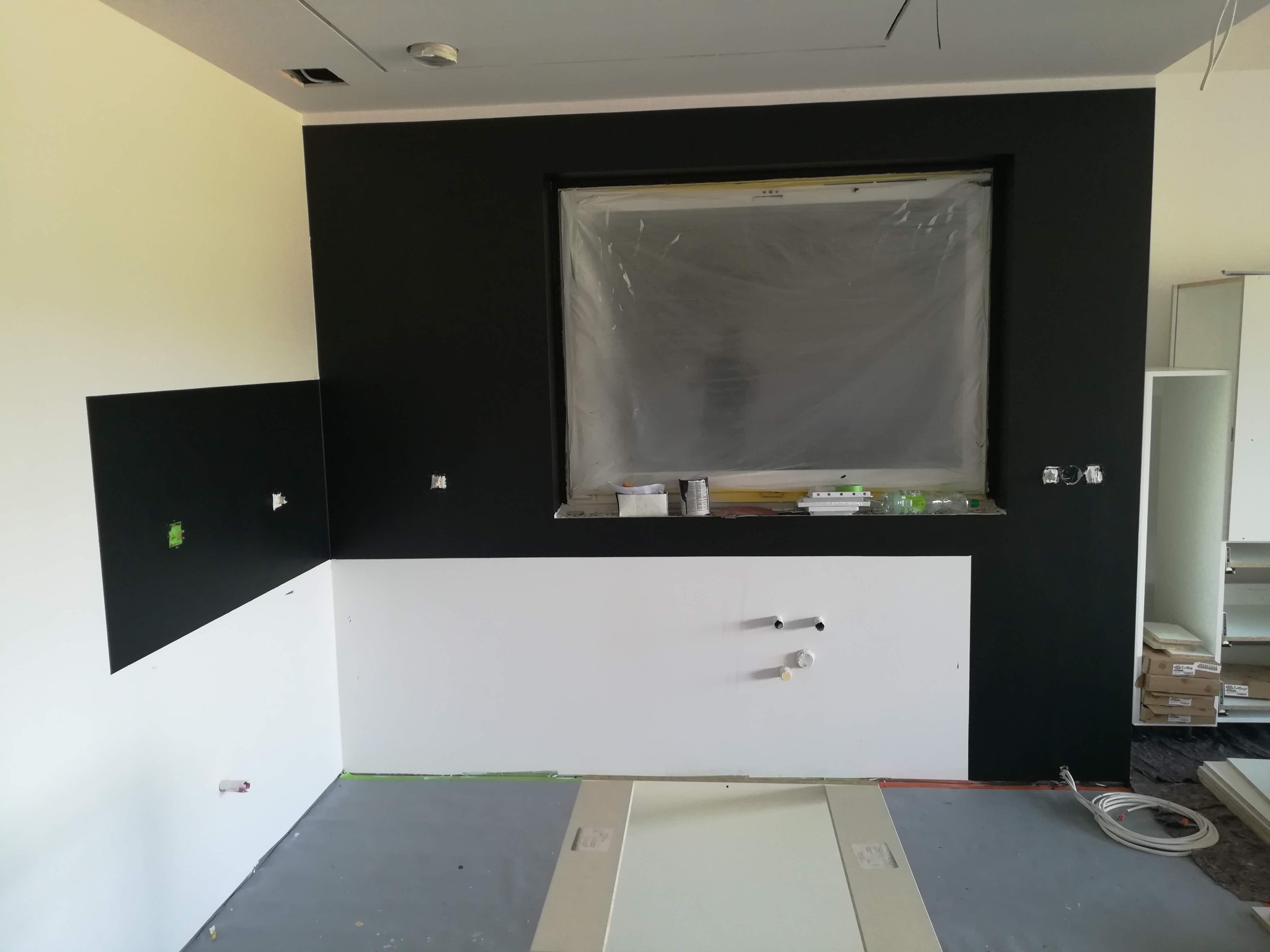 Wnętrze w trakcie malowania ścian na czarno i biało, okno zabezpieczone folią, widoczne puszki farb i narzędzia, przygotowane panele podłogowe.