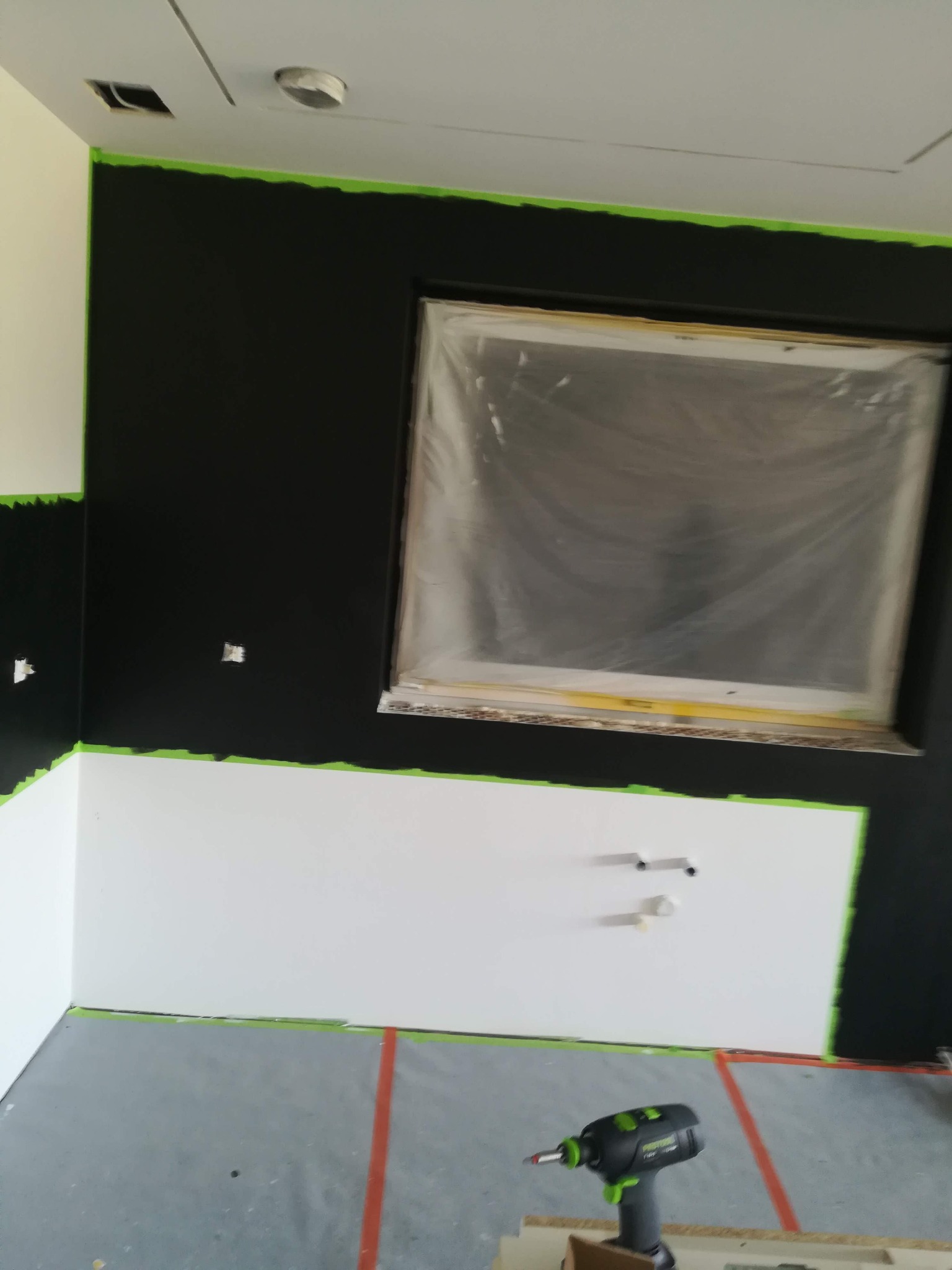 Ściana dwukolorowa, czarno-biała, zabezpieczona taśmą malarską, z zakrytym folią oknem i widocznym wiertarko-wkrętakiem na podłodze.