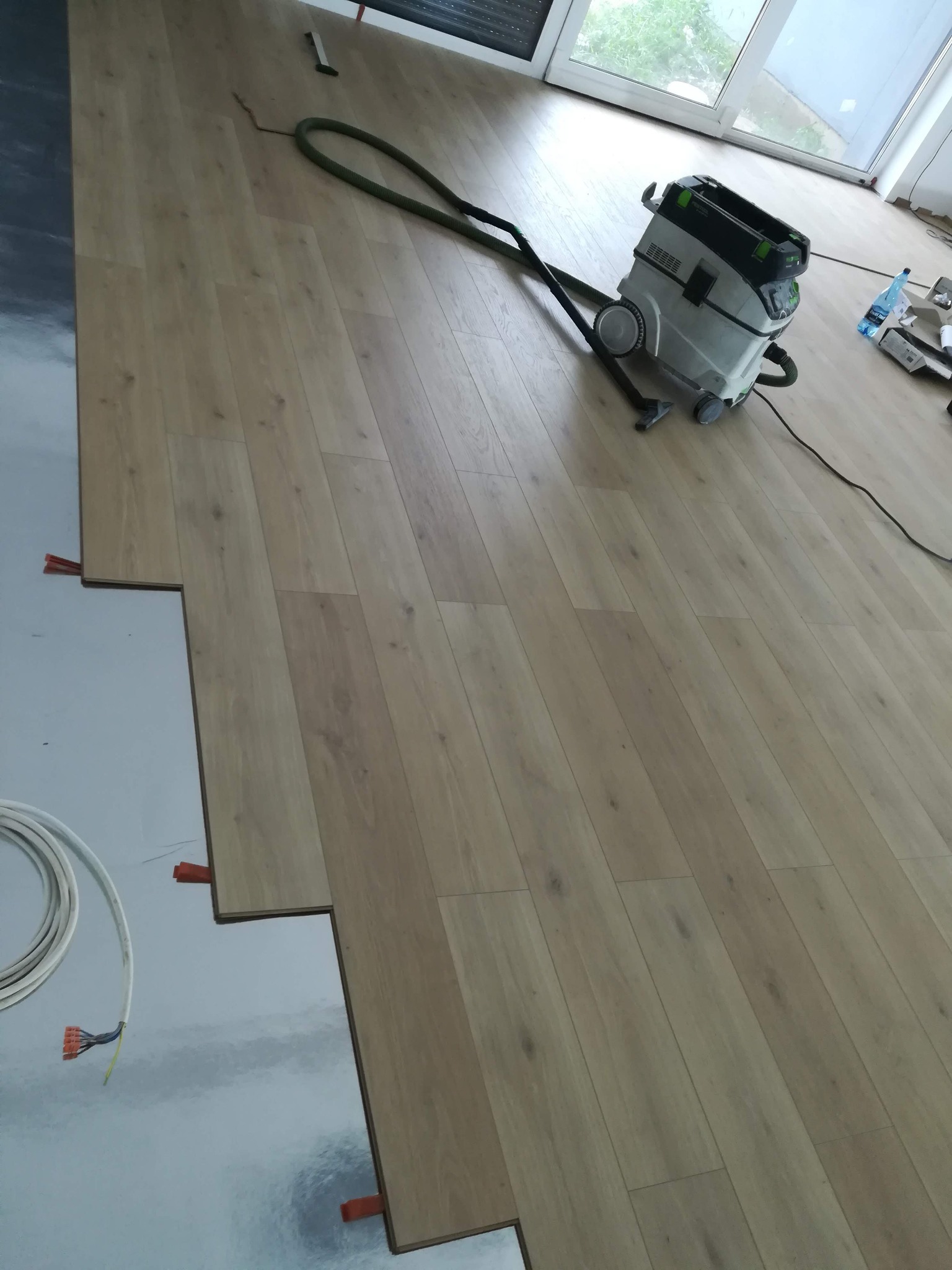 Proces układania paneli podłogowych z widocznymi dystansami, folią izolacyjną, kablem elektrycznym i odkurzaczem przemysłowym Festool na tle okna.