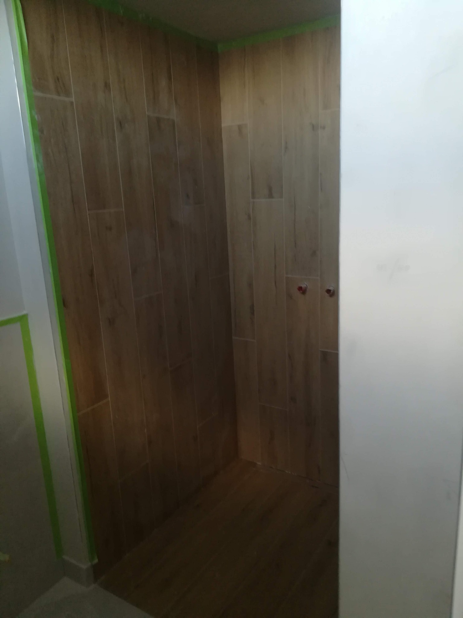 Wykończona kabina prysznicowa w Opolu z drewnopodobnymi płytkami, zabezpieczona taśmą malarską przy suficie i ścianach, z widocznymi przyłączami wodnymi.