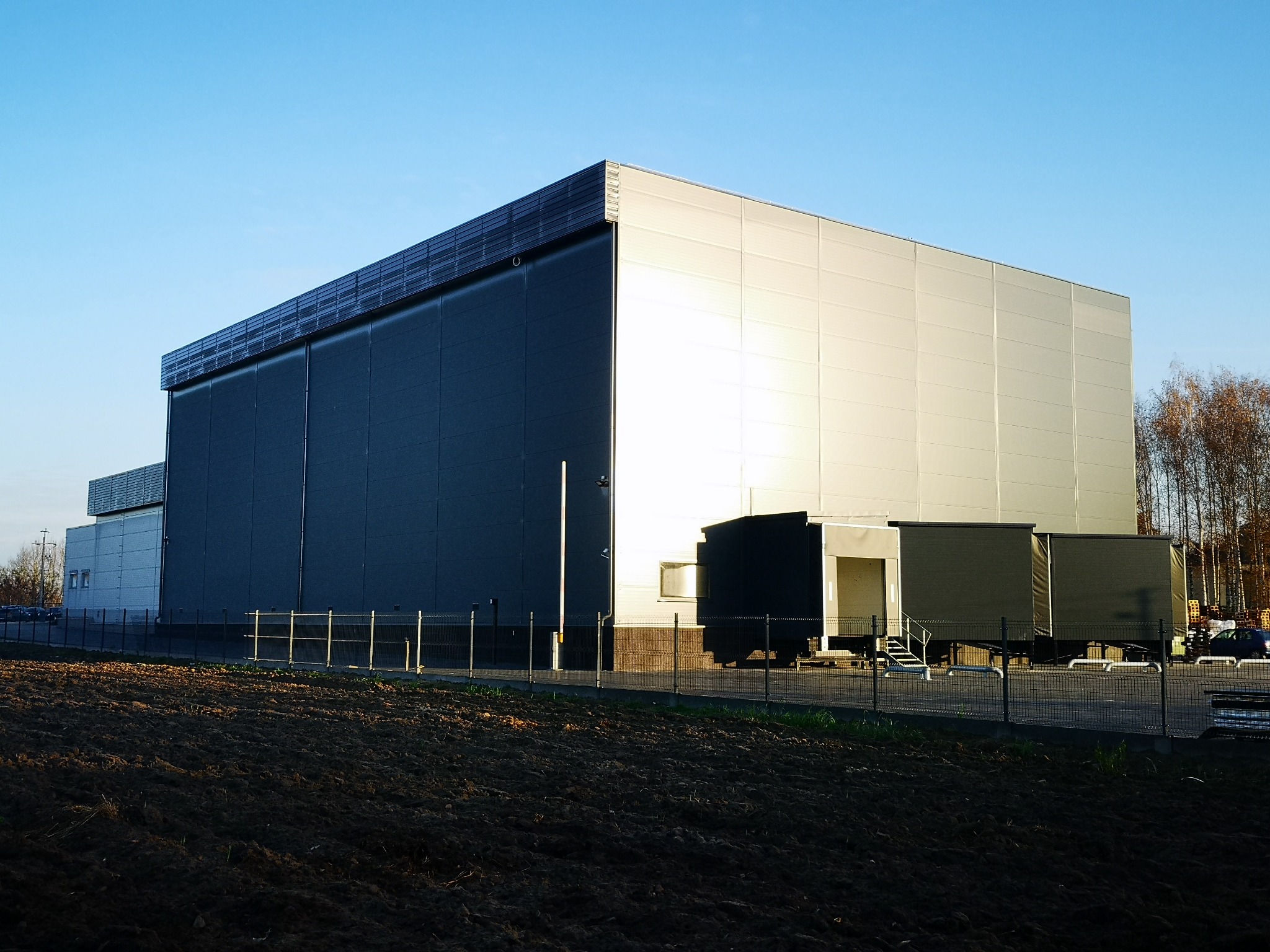 Duży, nowoczesny magazyn z panelami słonecznymi na dachu, z rampami rozładunkowymi i zaparkowanymi ciężarówkami, widziany z pola w słoneczny dzień.