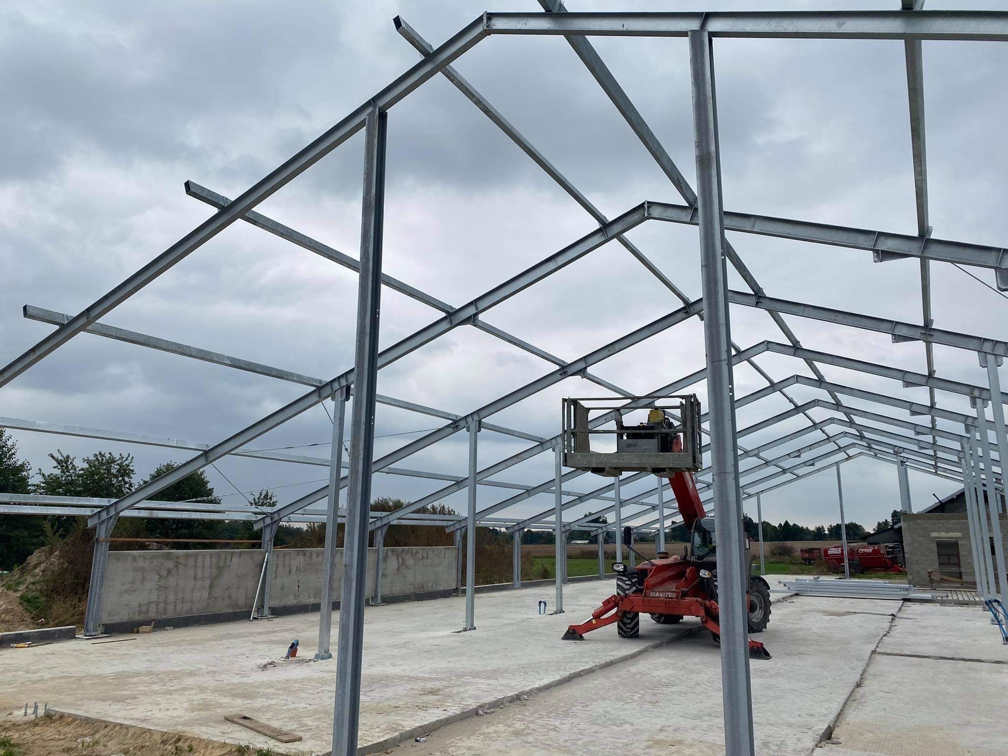 Montaż stalowej konstrukcji hali z użyciem podnośnika teleskopowego Manitou na betonowej płycie fundamentowej, widoczne elementy konstrukcyjne i pochmurne niebo.