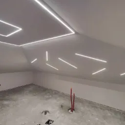 Poddasze w trakcie wykańczania z nowoczesnym oświetleniem LED na suficie, widoczne przewody instalacji elektrycznej wystające z podłogi.